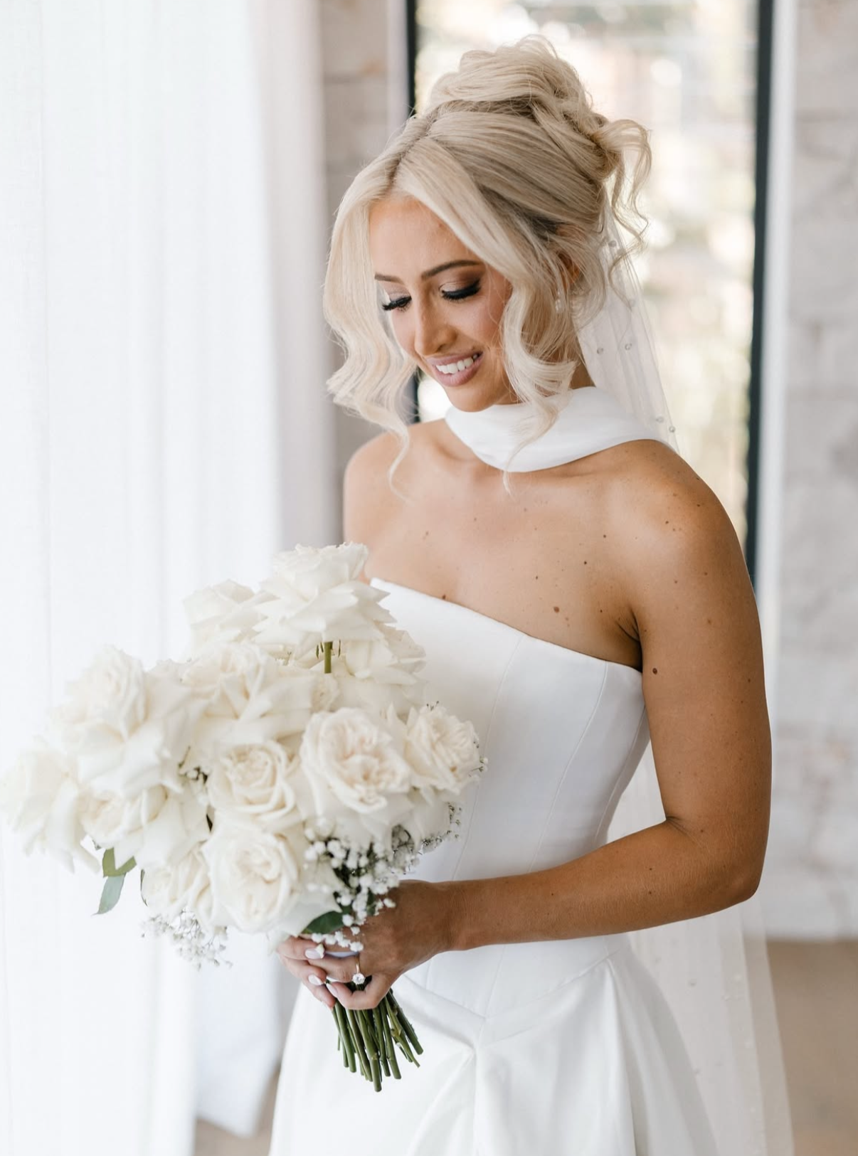 A bride with blonde hair styled in loose curls, wearing a strapless white wedding gown, is holding a bouquet of white roses and other flowers, smiling softly while looking down.