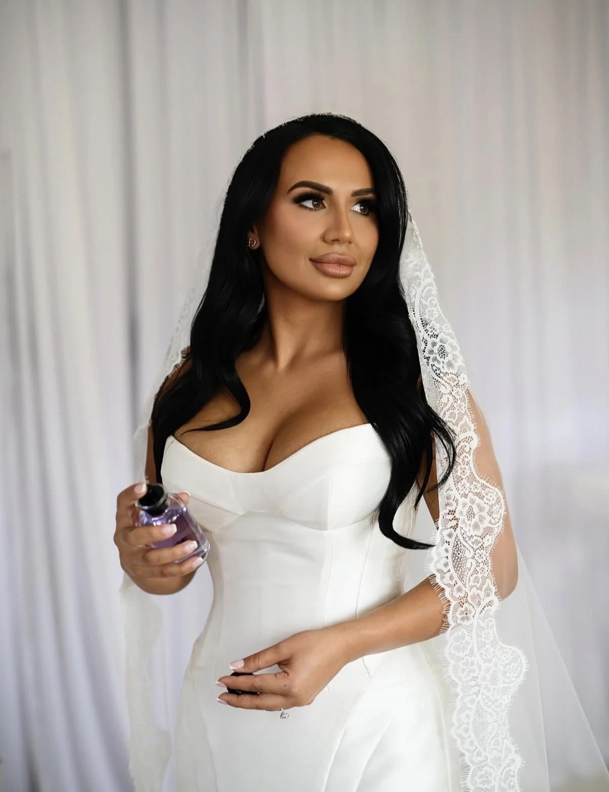 A woman in a white strapless wedding dress, holding a small perfume bottle, with long dark hair and a lace veil.