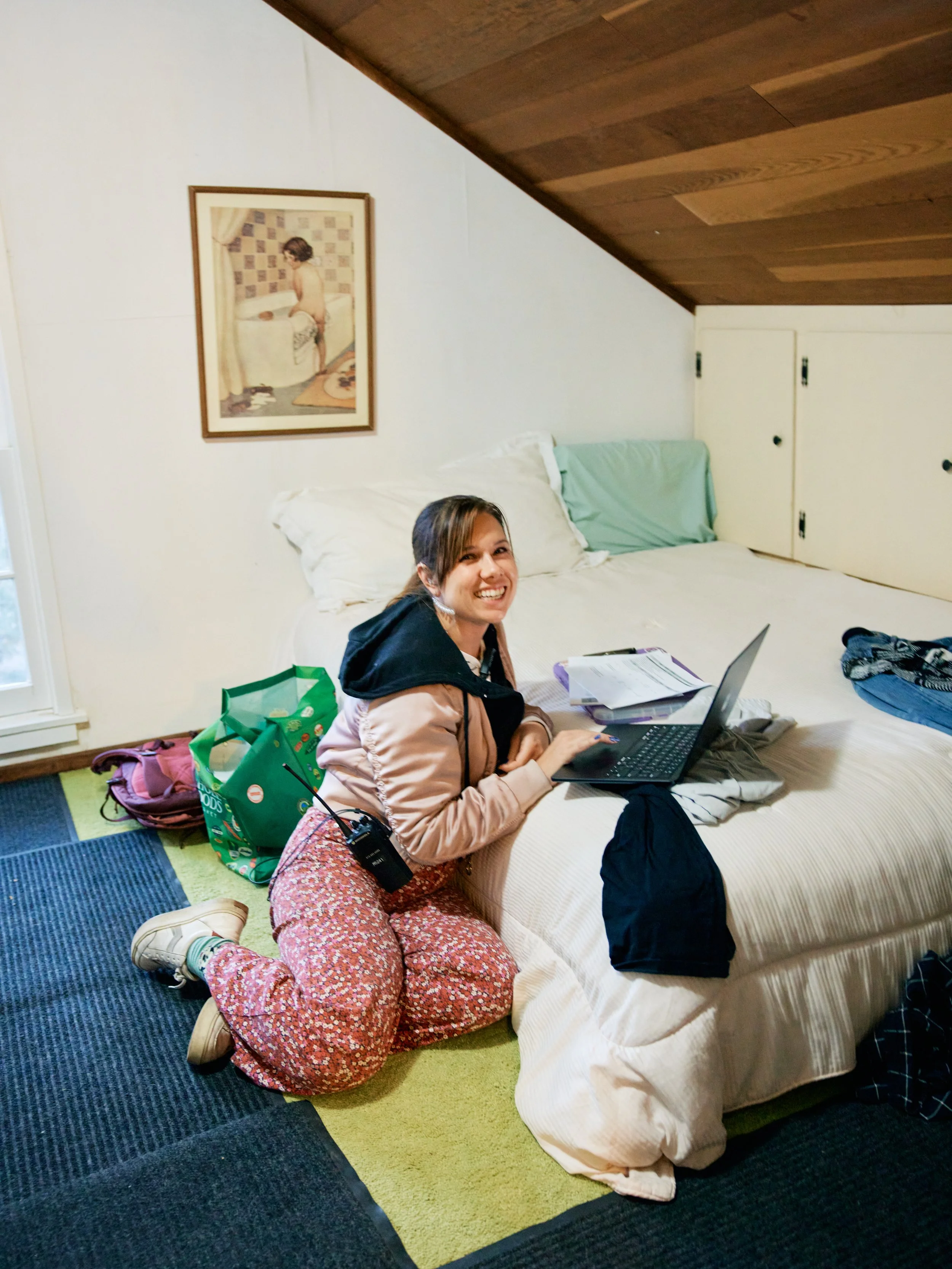 A young woman with brown hair, glasses, and a light pink jacket is sitting on the floor next to a bed, smiling at the camera. She has a walkie-talkie clipped to her pants. There are bags and school supplies around her, and she is wearing sneakers and floral-patterned pants. The bed has white sheets and pillows, and the wall behind shows a framed painting of a boy in a room with a bed and curtain. The room has a green rug, the floor has blue, green, and black carpeting, and there is a window with white curtains.