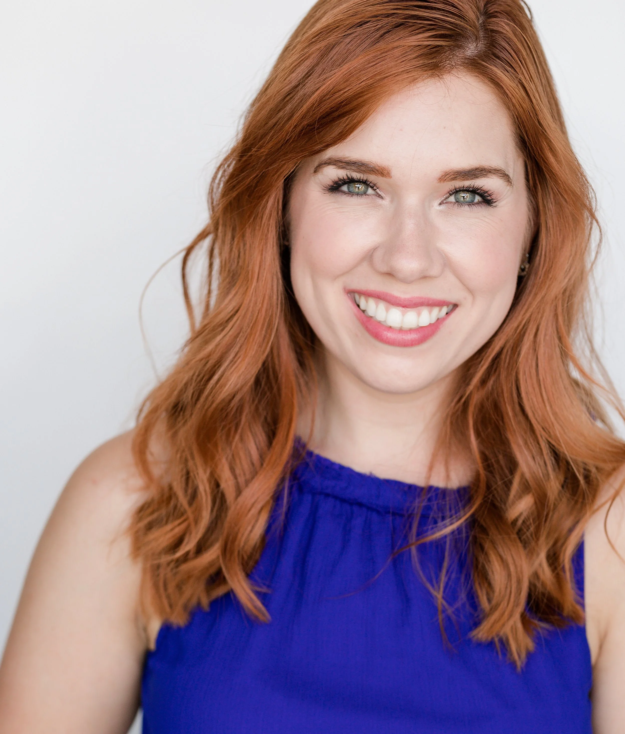Close-up of a smiling woman with red hair, blue eyes, light makeup, wearing a blue sleeveless top, against a plain white background.