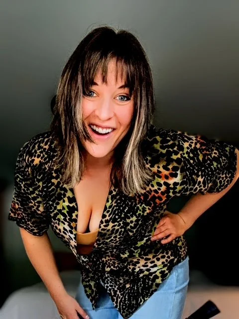 A woman with shoulder-length dark hair with highlights, smiling and leaning forward slightly, wearing a leopard print blouse and blue jeans.