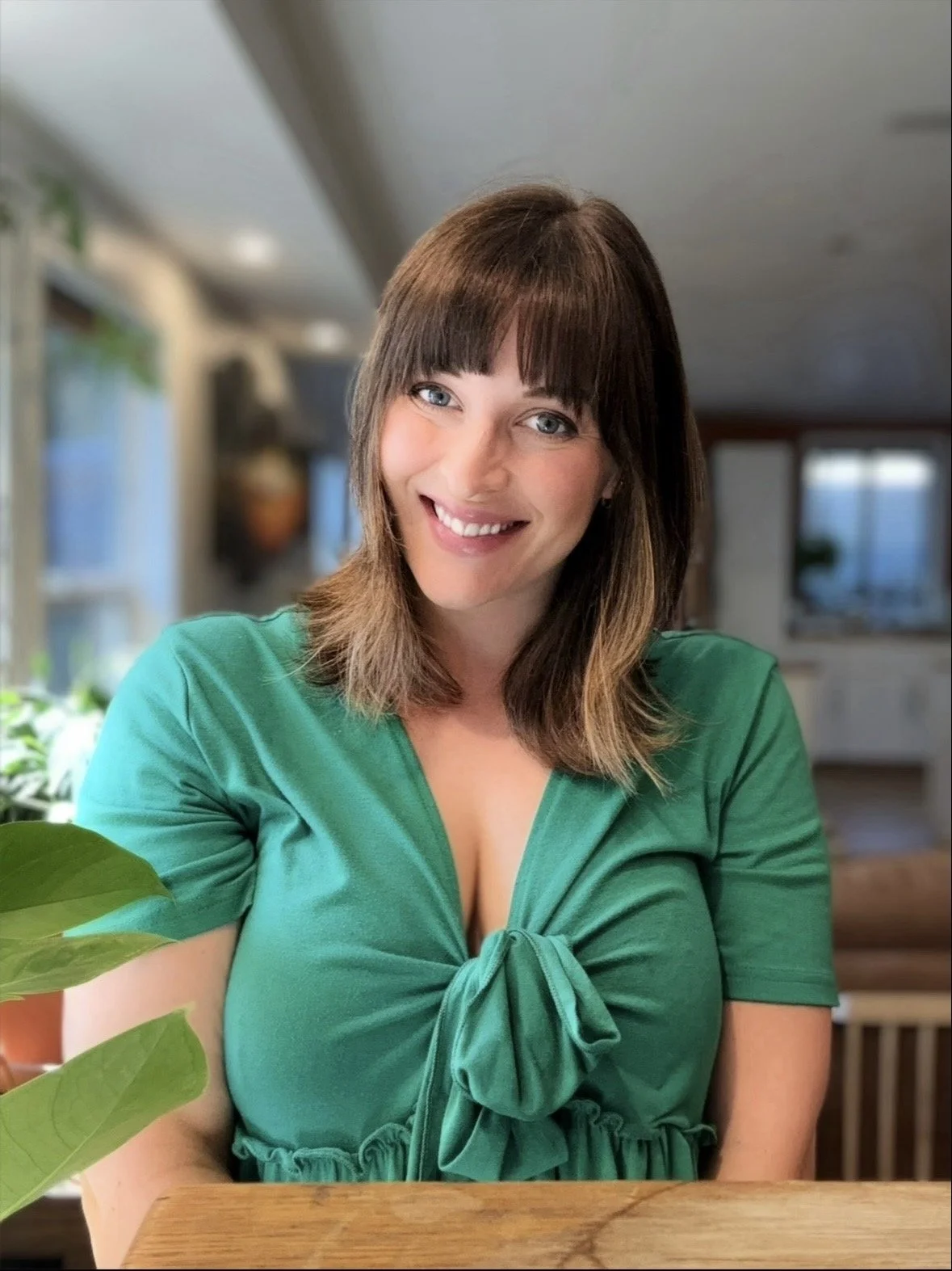 A woman with shoulder-length brown hair and blue eyes, smiling, wearing a green top with a bow at the front, sitting at a wooden table in a well-lit room with windows and plants.