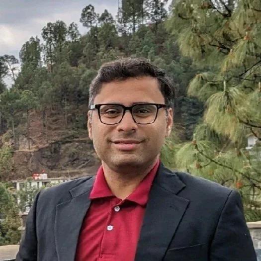 A man in glasses wearing a red shirt and dark blazer stands outdoors in front of a forested hillside with some trees visible.