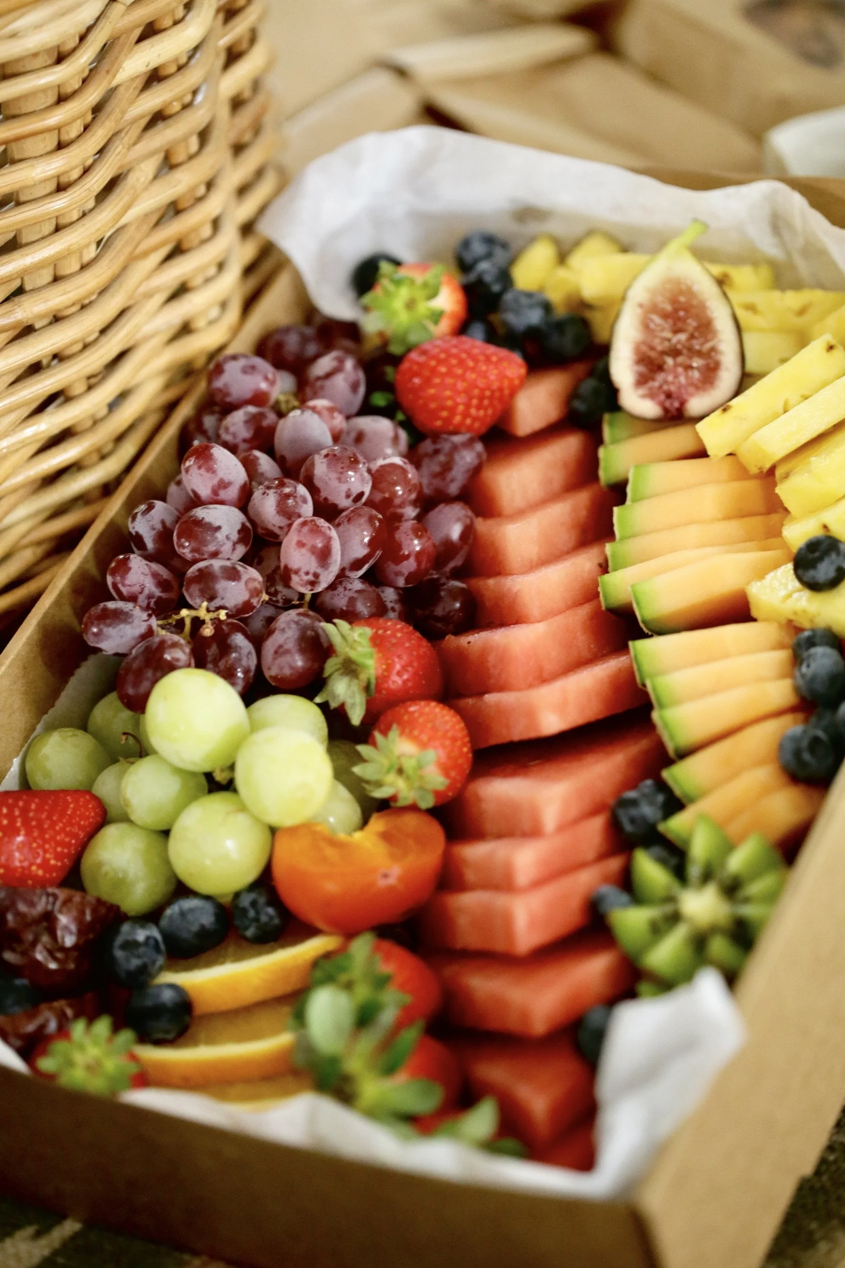 Assorted sliced fresh fruits including grapes, strawberries, pineapples, watermelon, blueberries, figs, and melon, arranged in a basket.