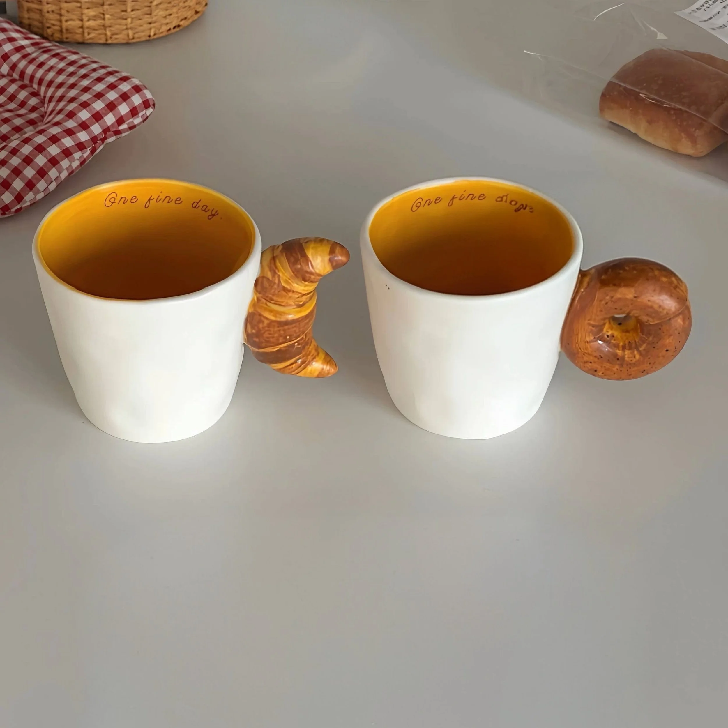 Cute ceramic mugs with croissant and bagel-shaped handles, featuring warm buttery tones and playful bakery-inspired design for cosy coffee moments.