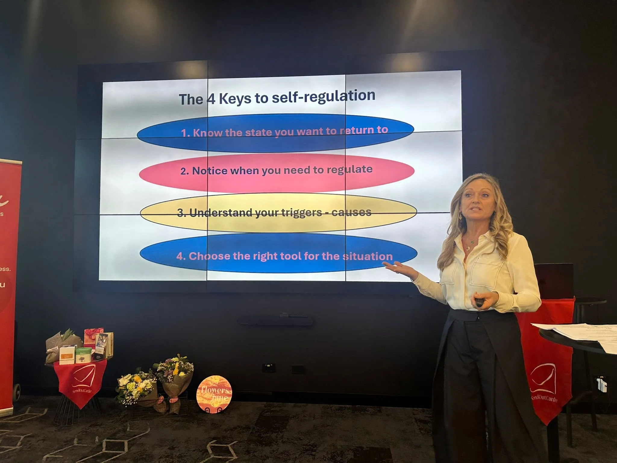 A woman standing in front of a presentation screen showing four keys to self-regulation. The keys are listed as: 1. Know the state you want to return to, 2. Notice when you need to regulate, 3. Understand your triggers - causes, 4. Choose the right tool for the situation. She is gesturing with her left hand and appears to be giving a presentation. There are flowers and gift bags on a table below the screen.