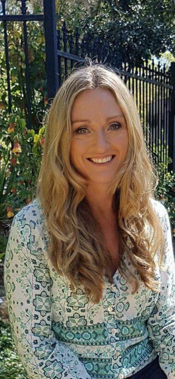 A woman smiling outdoors with long, curly blonde hair, wearing a patterned blouse, in front of greenery and a black metal fence.