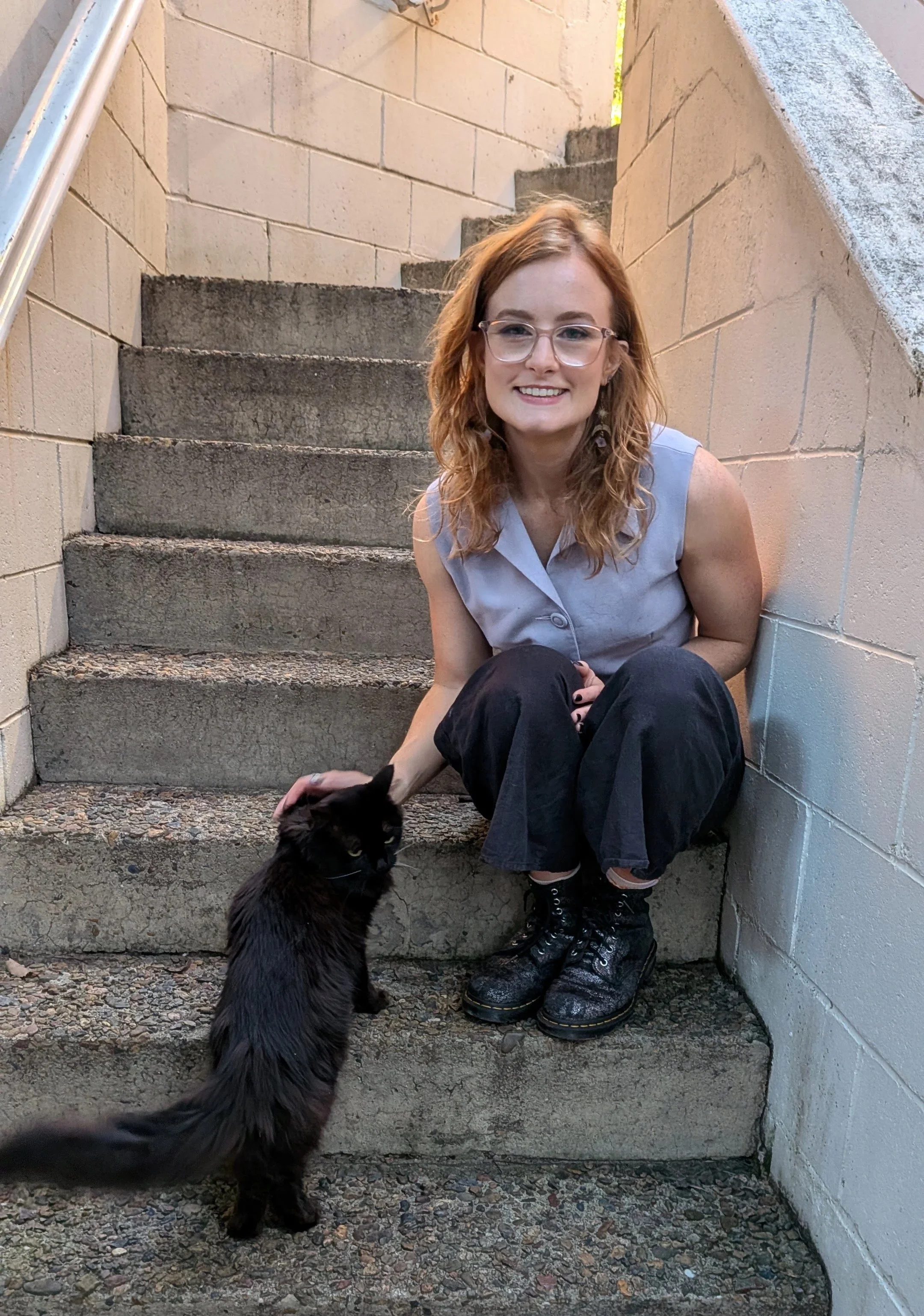 Phoebe sits on concrete steps smiling while gently petting a fluffy black cat approaching from the step below. Phoebe wears clear-framed glasses, a light blue sleeveless vest, loose black pants and sparkly black boots.