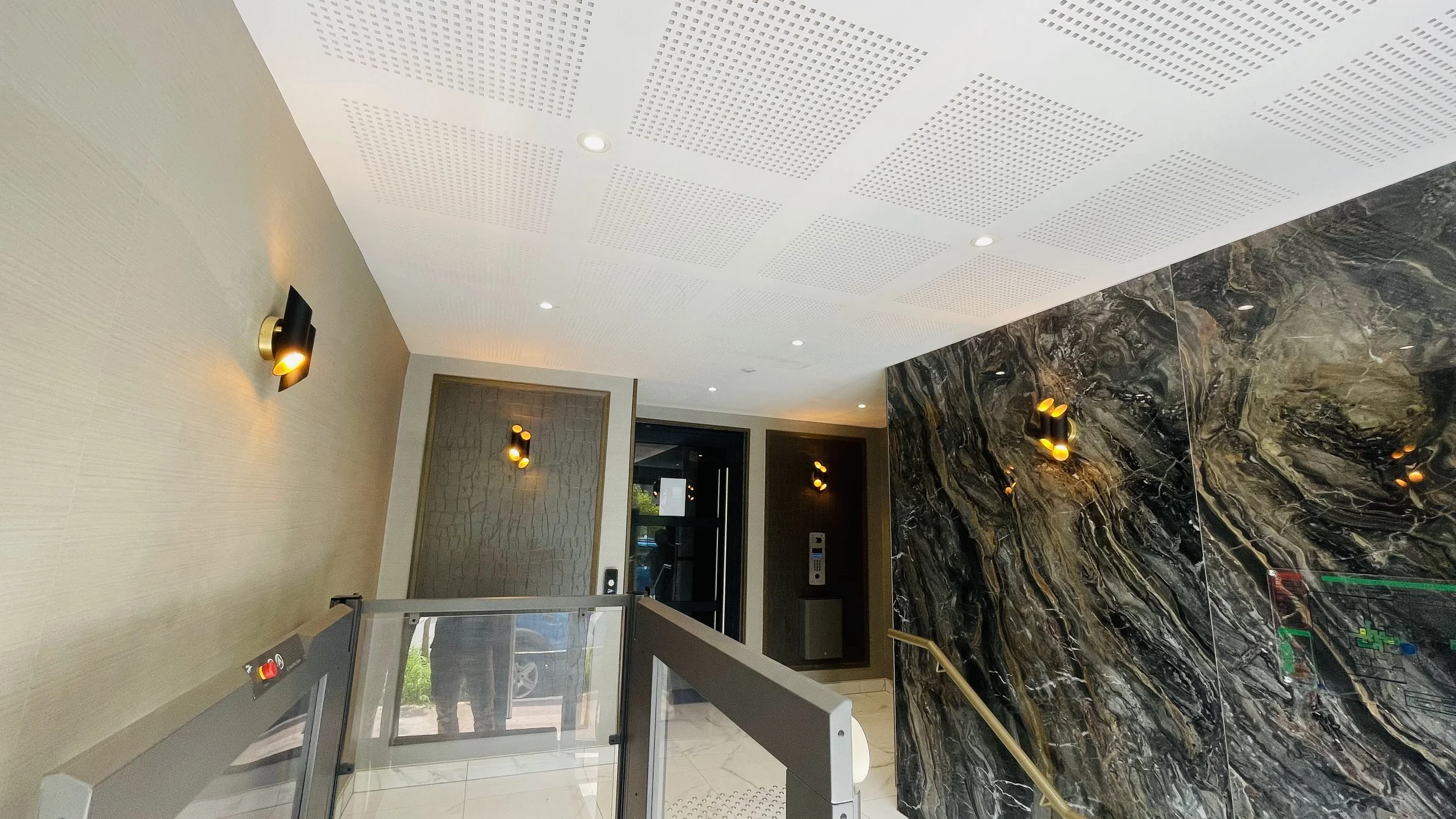 Intérieur d'un bâtiment avec un plafond blanc à motifs perforés, des murs en marbre noir avec des veines dorées, et des appliques murales en forme de projecteurs noirs avec lumière jaune. On voit également une porte en verre à l'arrière et un ascenseur avec un bouton d'appel près de l'entrée.