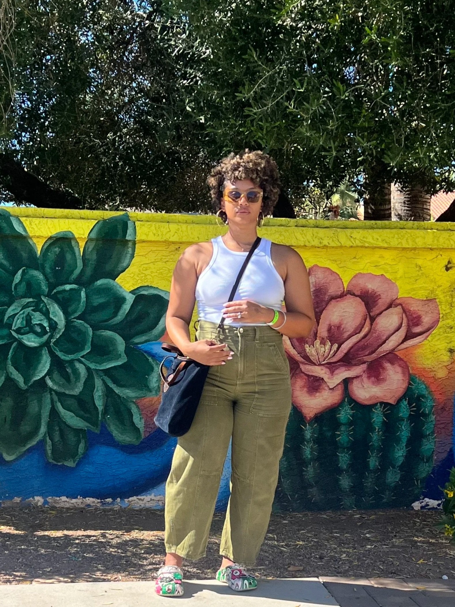 Afro-Latina, black woman with natural hair, white tank top, green cargo barrel pants, and colorful crocs standing in front of a vibrant mural with large floral designs.