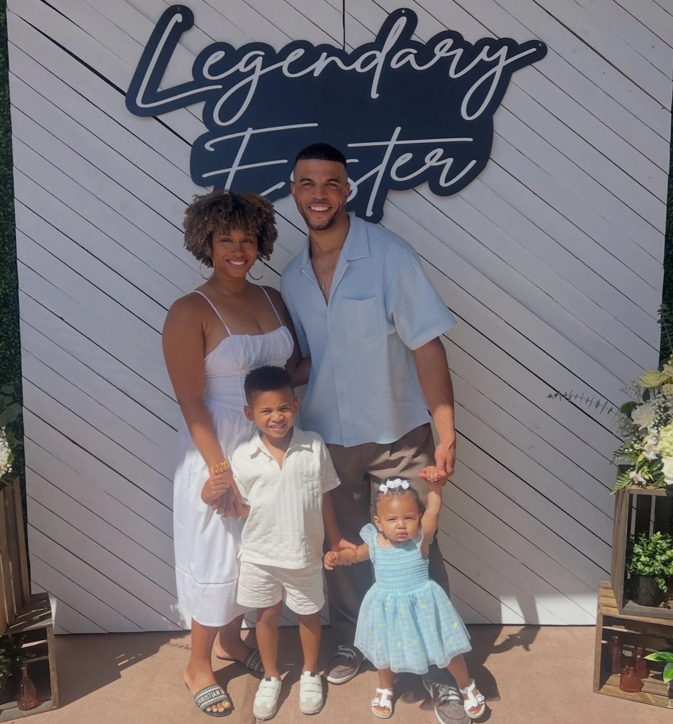 A family of five smiling and posing for a photo in front of a white wooden wall with a sign that says 'Legendary Easter.' The group includes a woman with curly hair, a man, a young boy, and two young girls, all dressed in light-colored summer clothes.