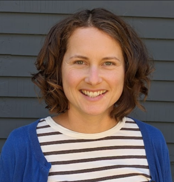A woman with curly brown hair, wearing a white and black striped shirt and blue cardigan, smiling in front of a gray wooden wall.
