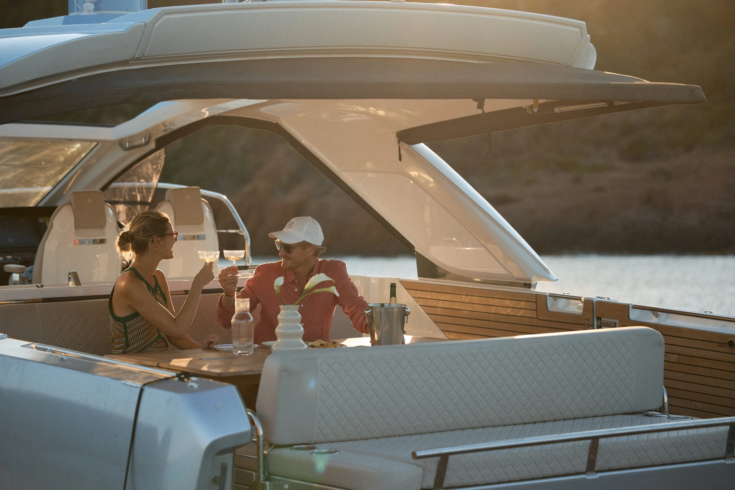 A man and woman having dinner on a yacht during sunset, raising glasses for a toast with a bottle of wine and table settings nearby.