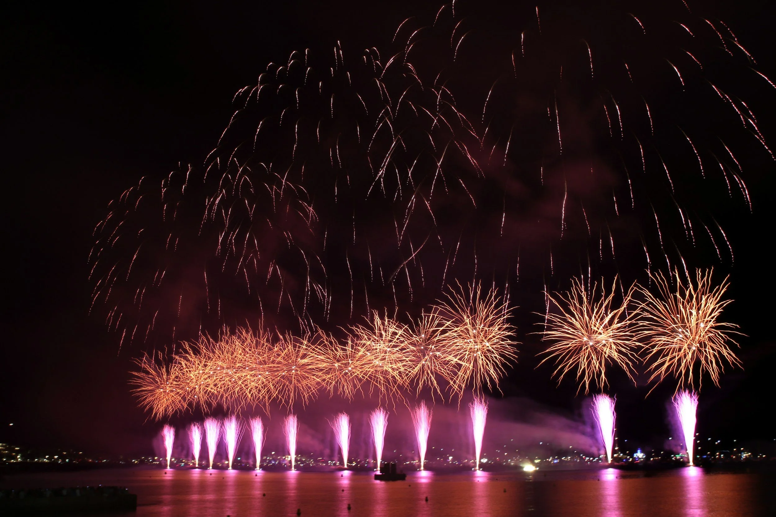 A fireworks display over a body of water at night with bursts of pink, purple, and orange lights reflecting on the water's surface.