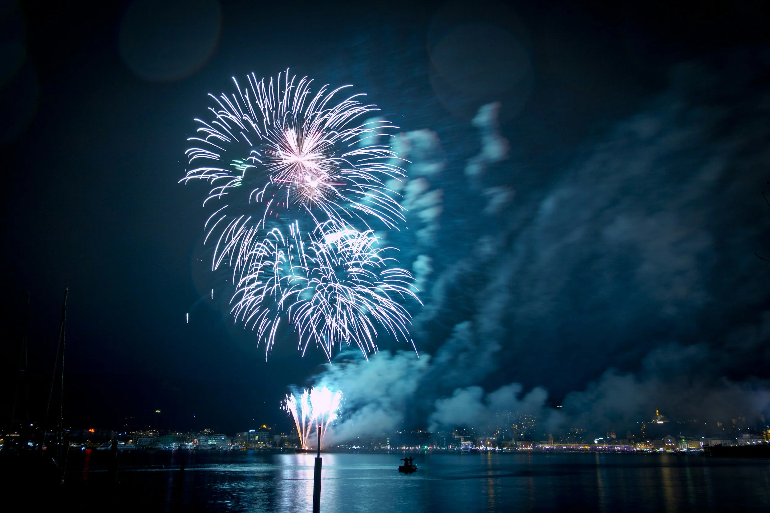 Fireworks display over a city waterfront at night, with reflections on the water and a boat in the foreground.