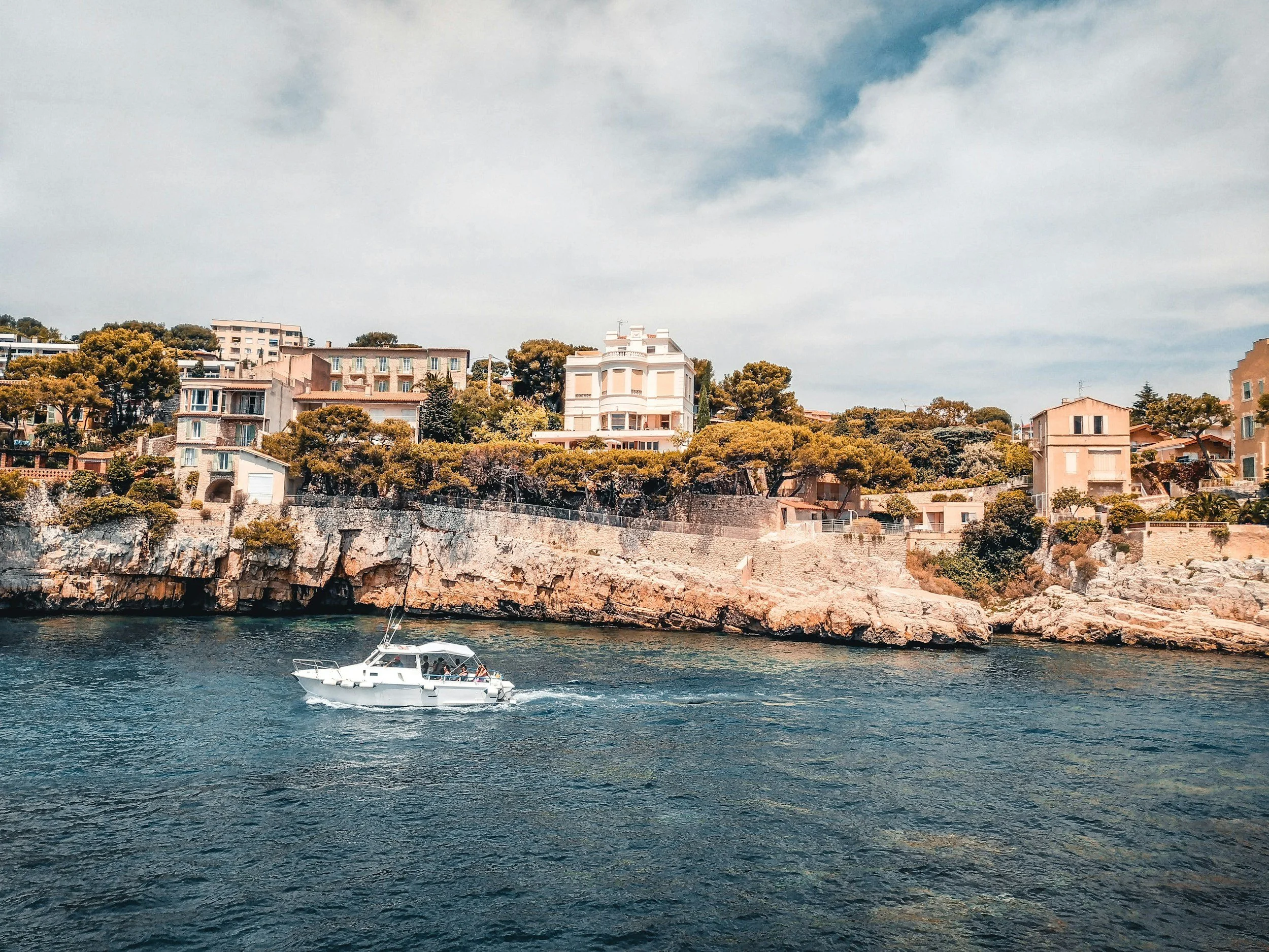 A boat sailing on a body of water with a rocky bank and a hillside dotted with residential buildings and trees in the background under a cloudy sky.