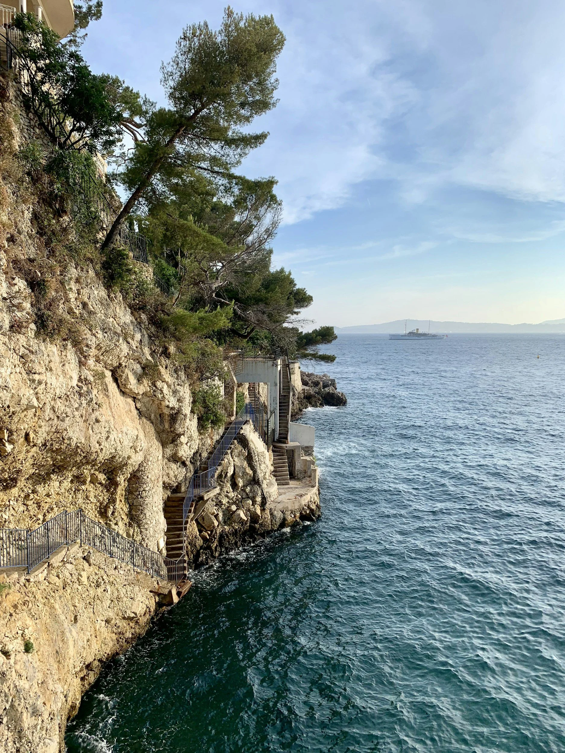 A rocky seaside cliff with steps and railings leading down to the water, trees growing along the edge, and a large ship sailing in the distance across a calm blue sea under a partly cloudy sky.