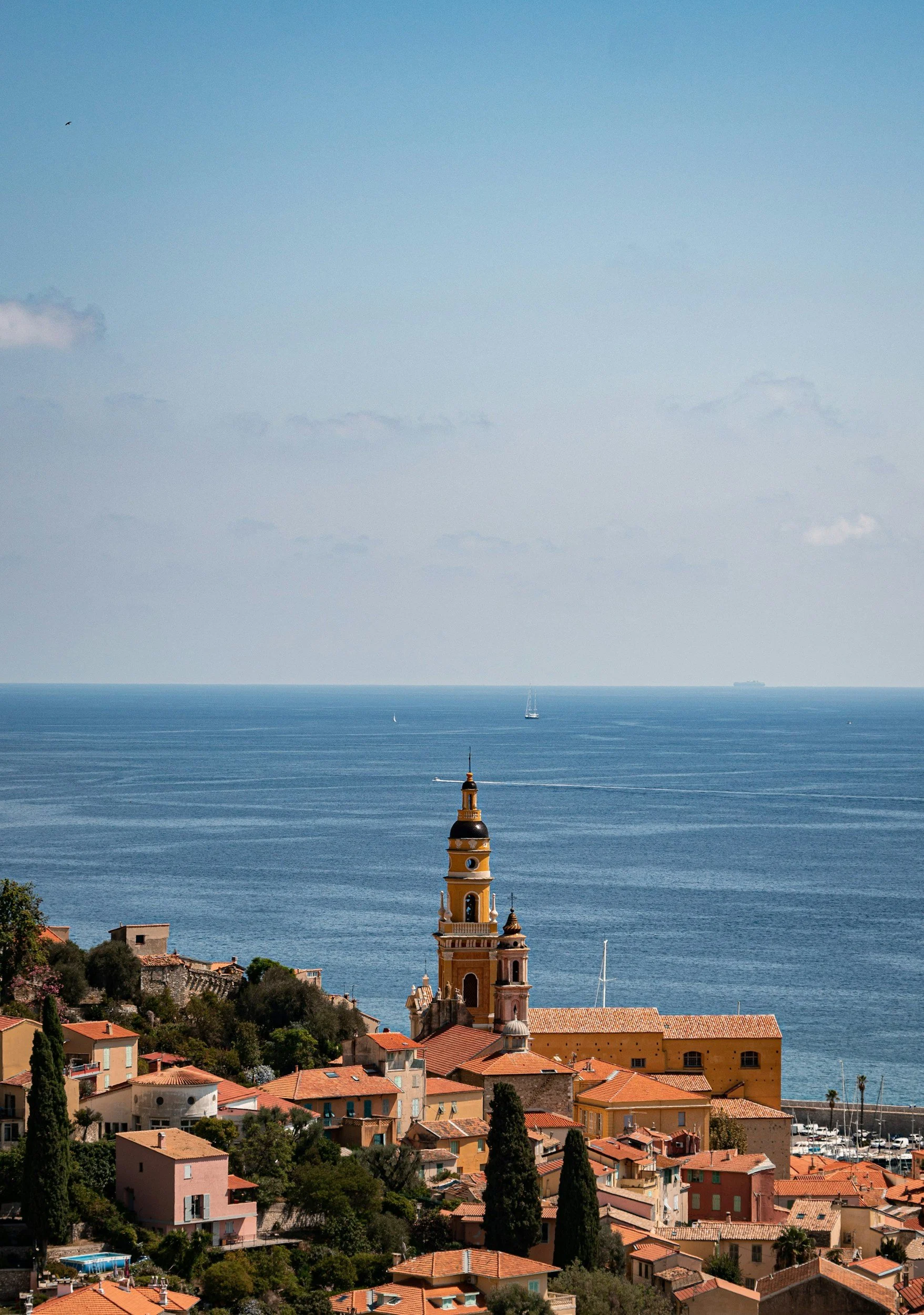 A scenic view of a coastal town with orange rooftops, a tall yellow and white church tower, green trees, and a calm blue sea with sailboats and a large cargo ship in the distance.