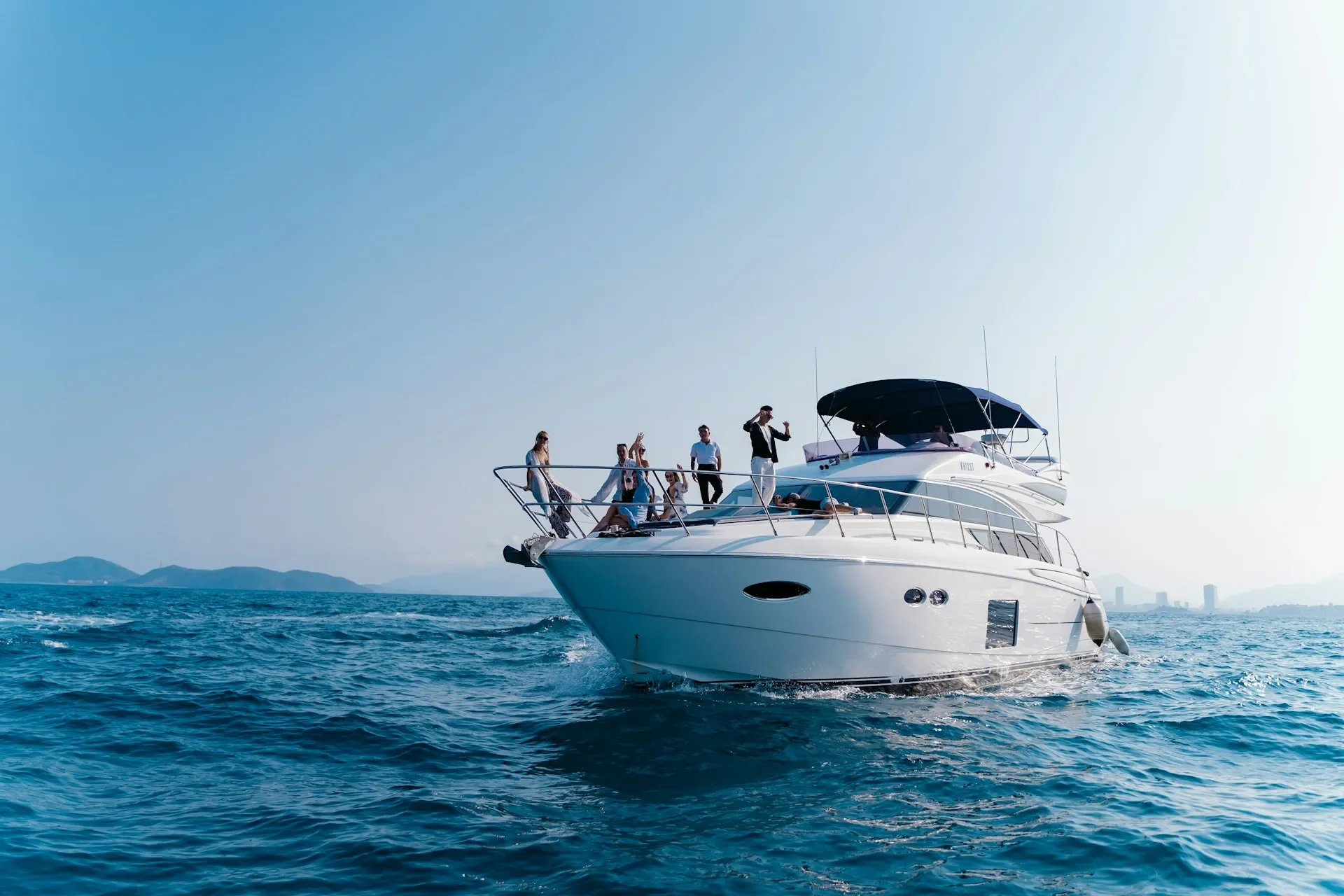 A white yacht with multiple people on board sailing in calm blue waters with city buildings and mountains in the background under a clear sky.