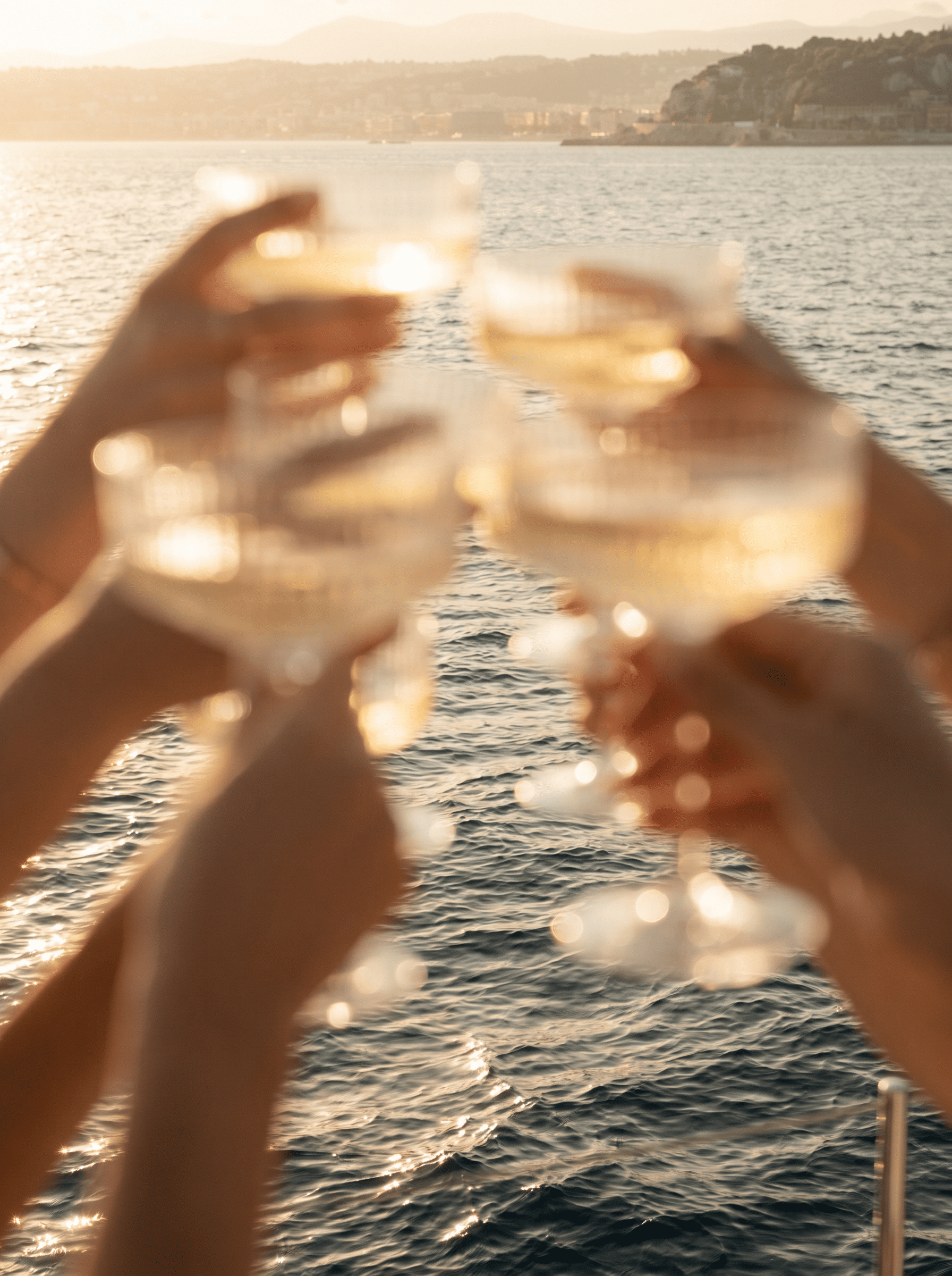 Two people holding glasses of white wine by a body of water during sunset.