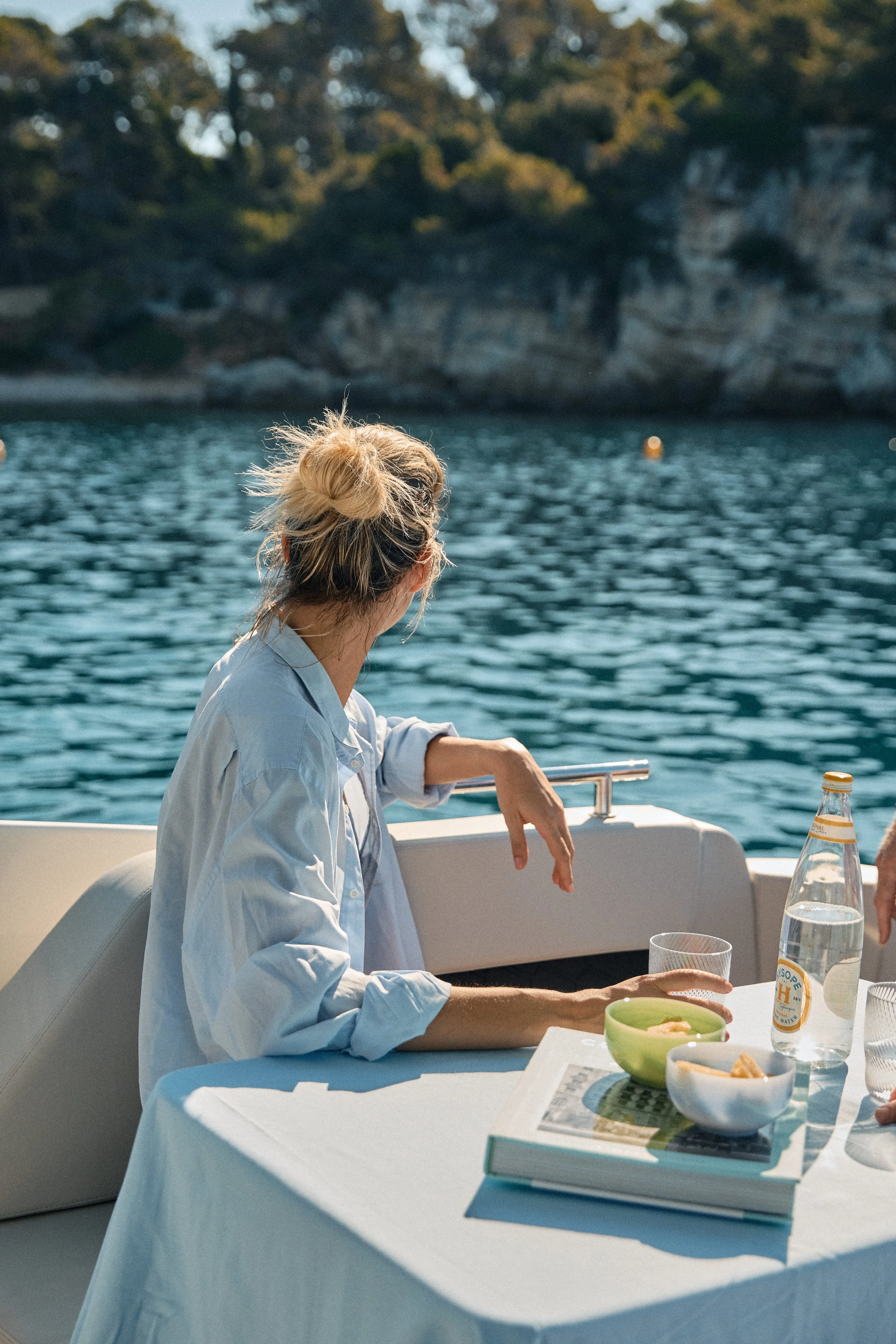 A woman with blonde hair tied in a bun, wearing a light blue shirt, sitting at a table on a boat. The table has a white tablecloth, a glass of water, a green bowl of food, and a book. The background shows water and trees.