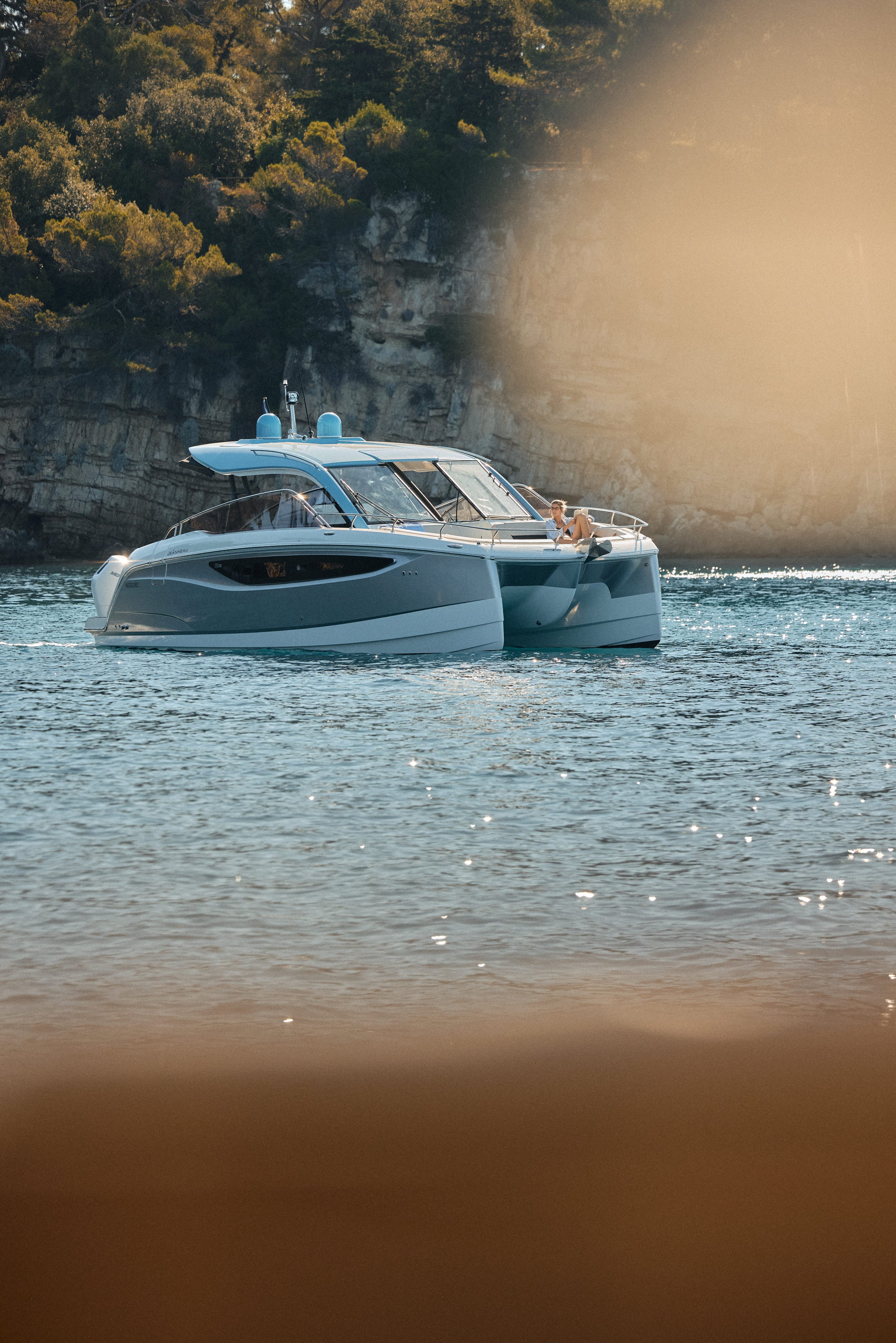 A white and blue yacht floating on calm water near rocky cliffs with trees, under a warm sunlight.