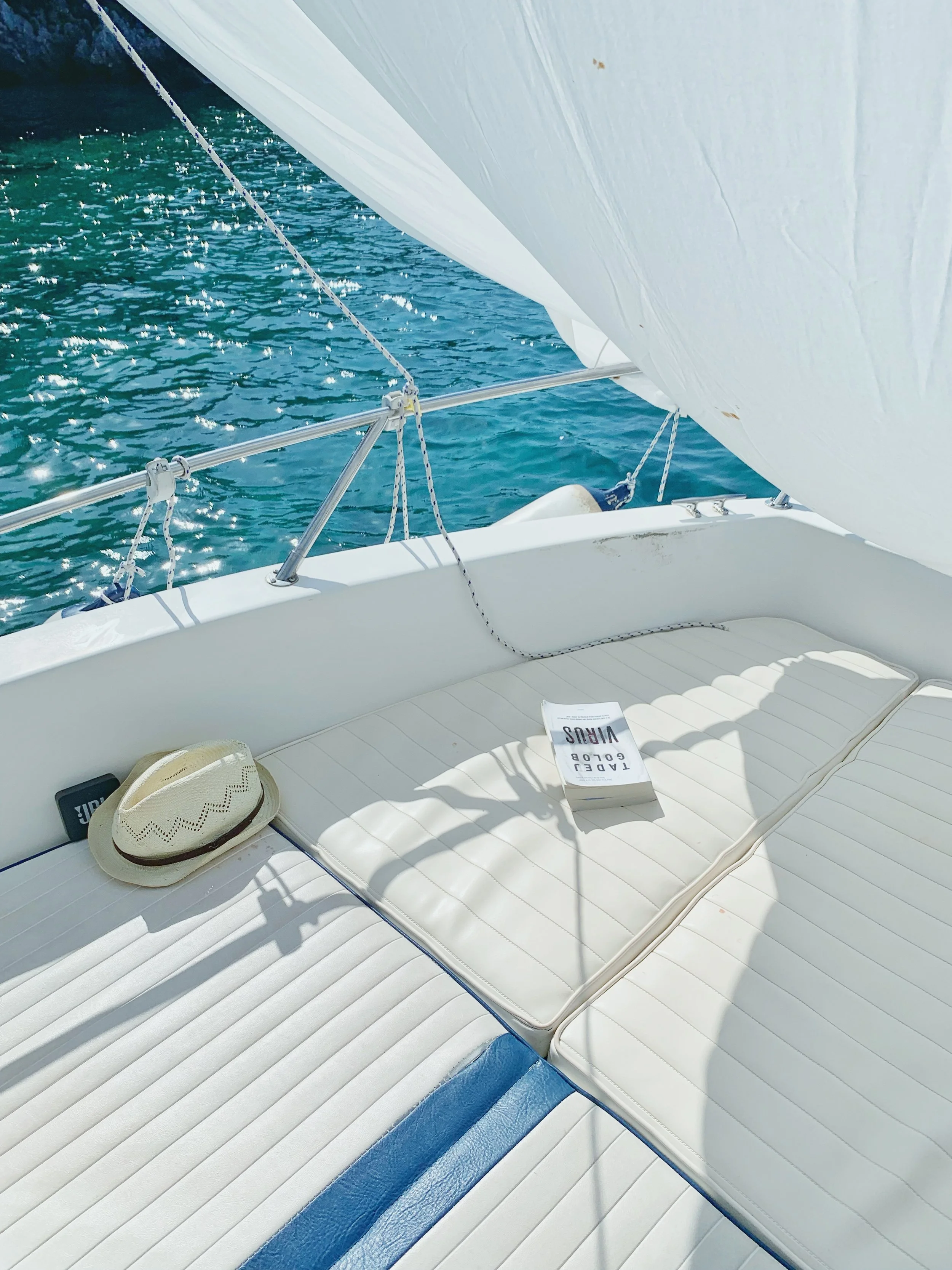 View from a yacht showing the boat's white cushioned deck, a straw hat, and a booklet, with sparkling blue water and part of the boat's sail in the background.
