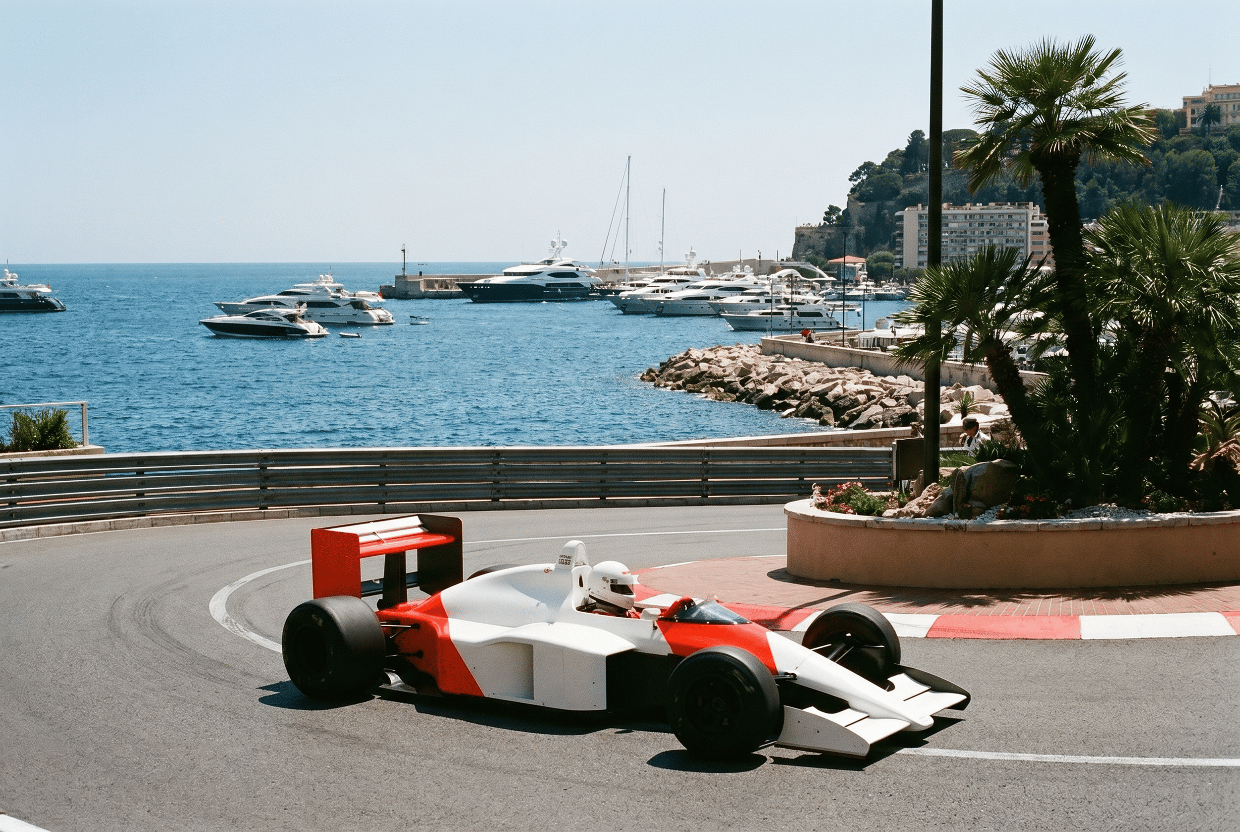 A red and white race car on a street near a harbor with yachts and sailboats docked, a palm tree, and buildings on a hill in the background.