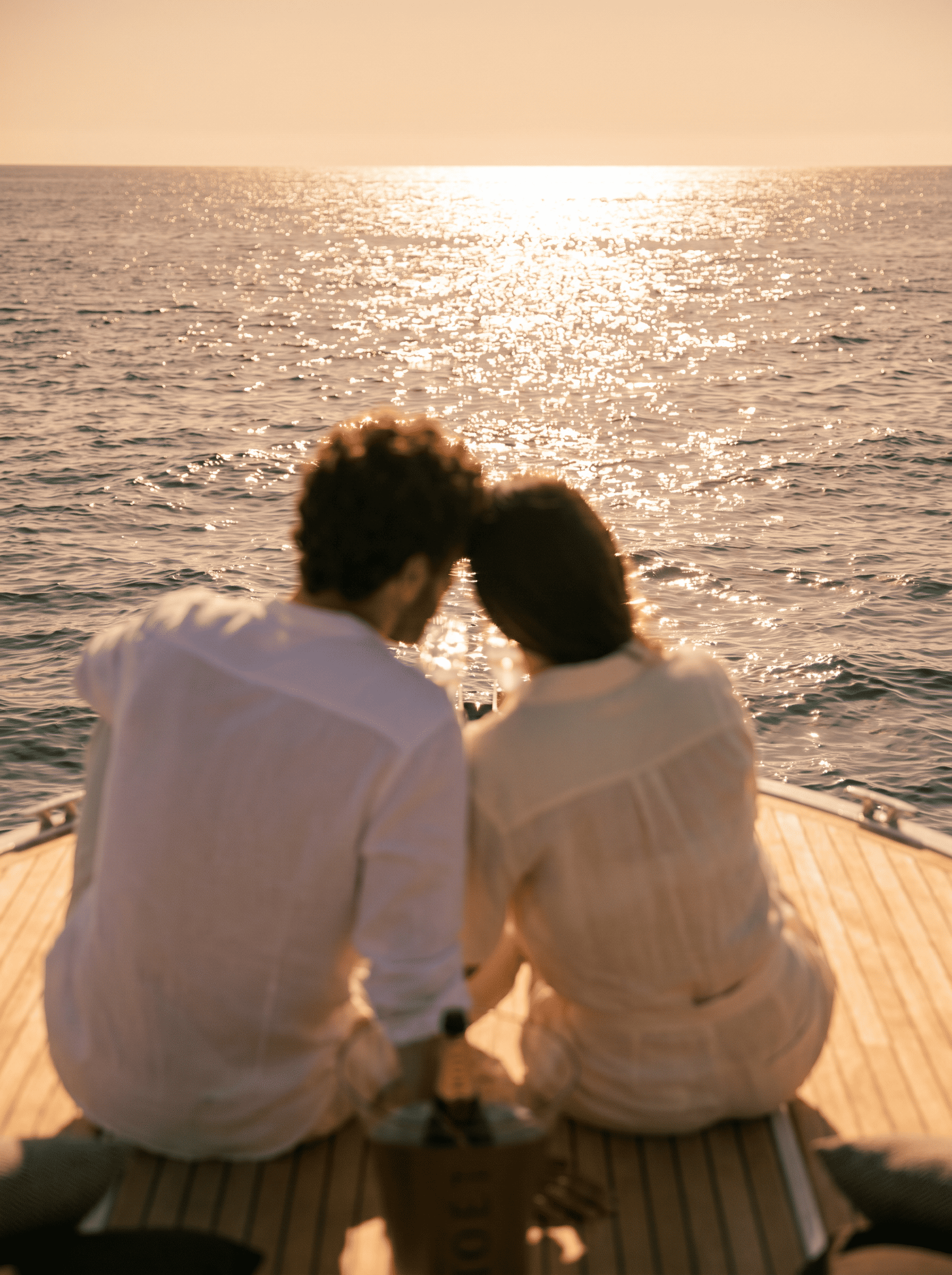 A couple is sitting close together on a boat, facing the water during sunset, with their heads touching.