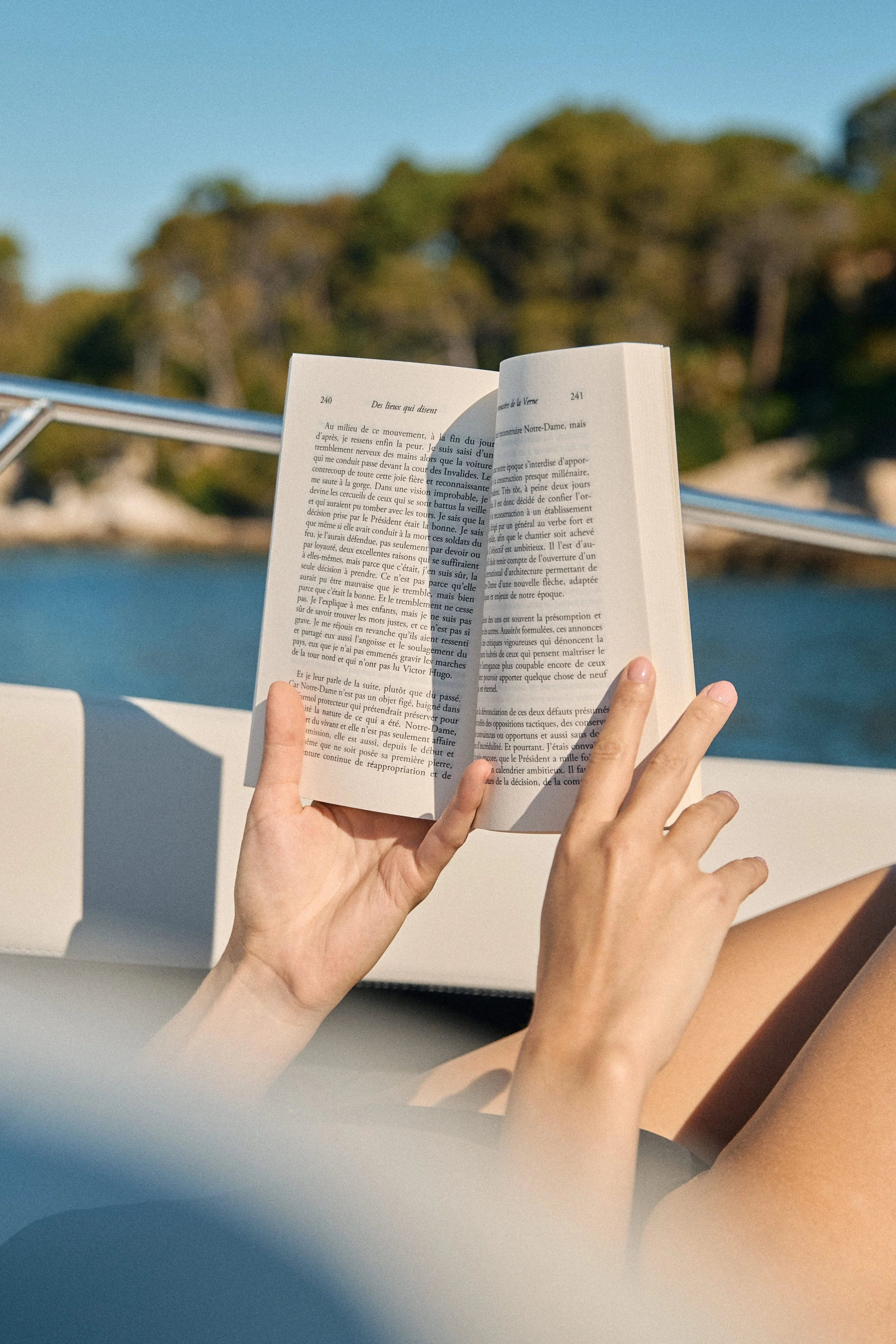 Person holding an open book with text in front of a boat with a blue railing, with trees and blue sky in the background.