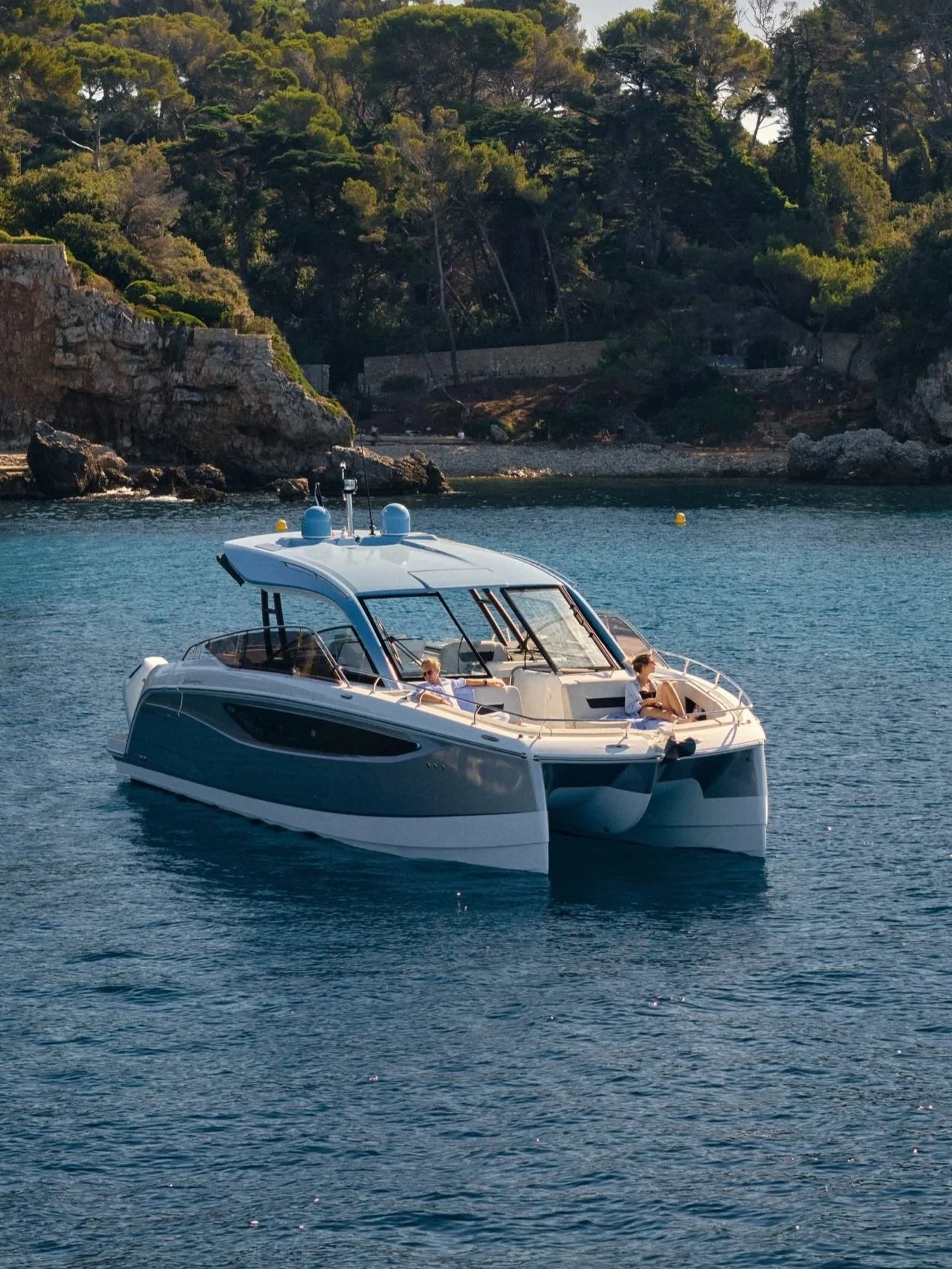 A white yacht with a black stripe on the water near a rocky shoreline with trees and a stone wall. Two people are relaxing on the deck of the yacht, enjoying the sunny day.