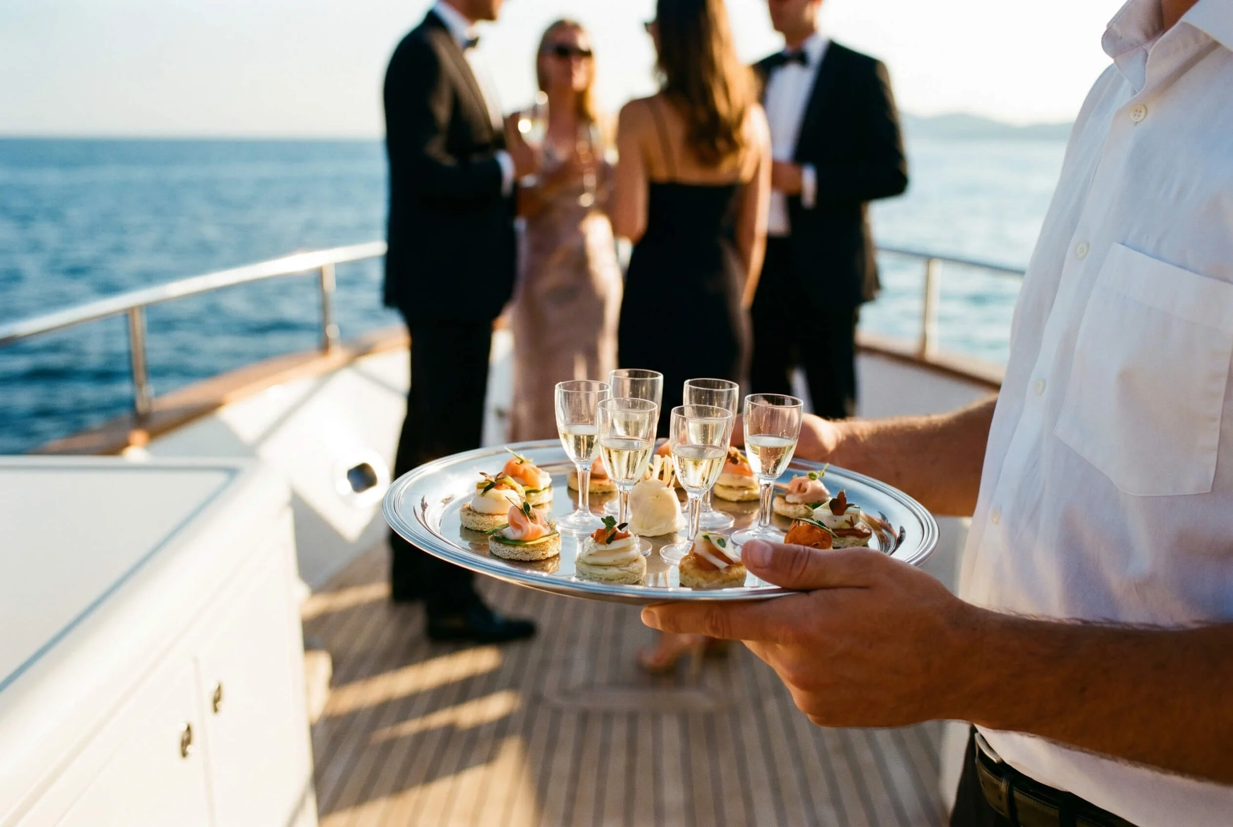 A person holding a silver tray with various small appetizers and glasses of champagne, with a blurred group of people in formal attire in the background on a boat.