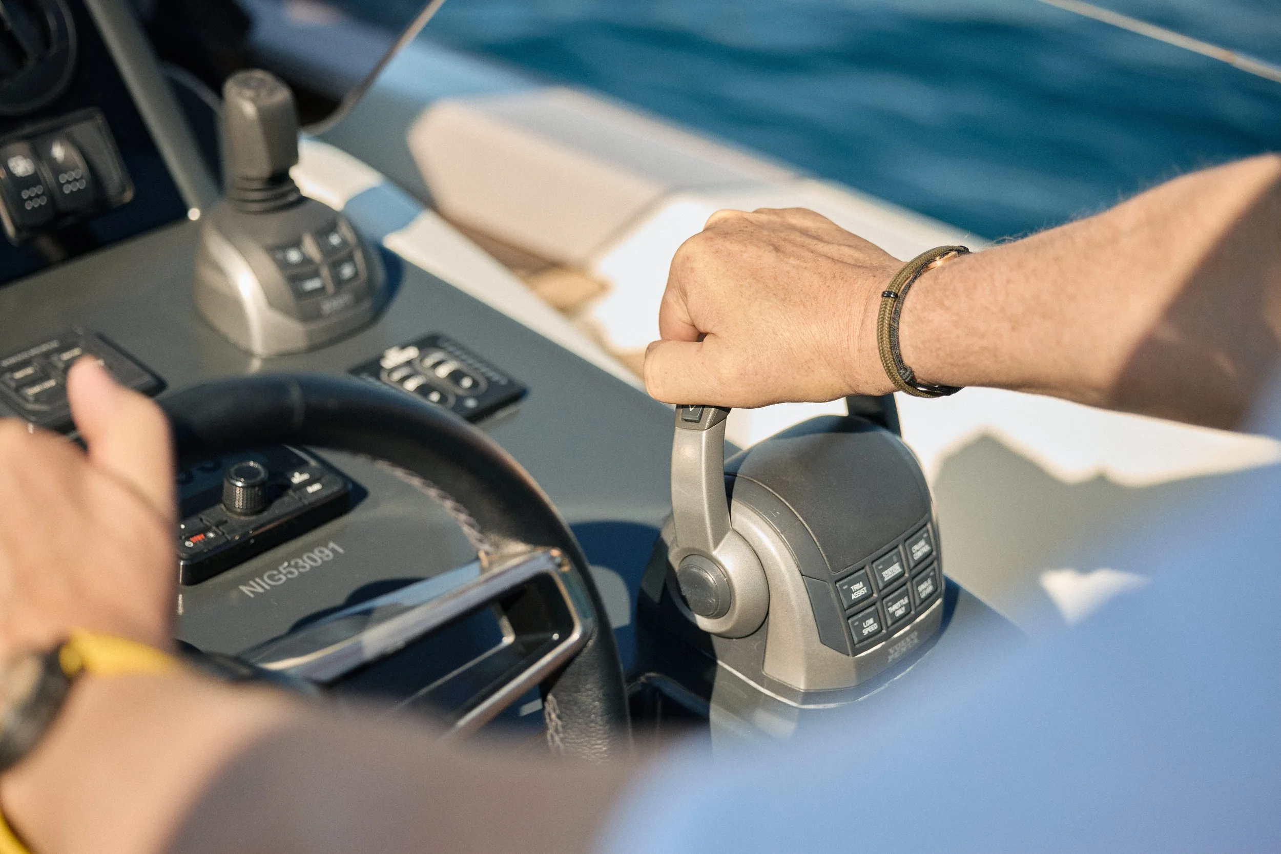 A person driving a boat, holding the throttle control with their right hand, wearing a bracelet, with a view of the boat's dashboard and water in the background.