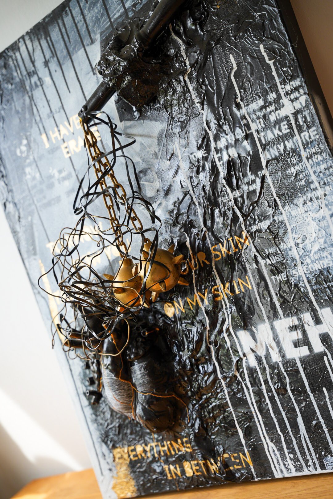 Mixed media art piece with black, gold, and metallic elements, featuring text and abstract textures, with layered materials including wires, chains, and paint.