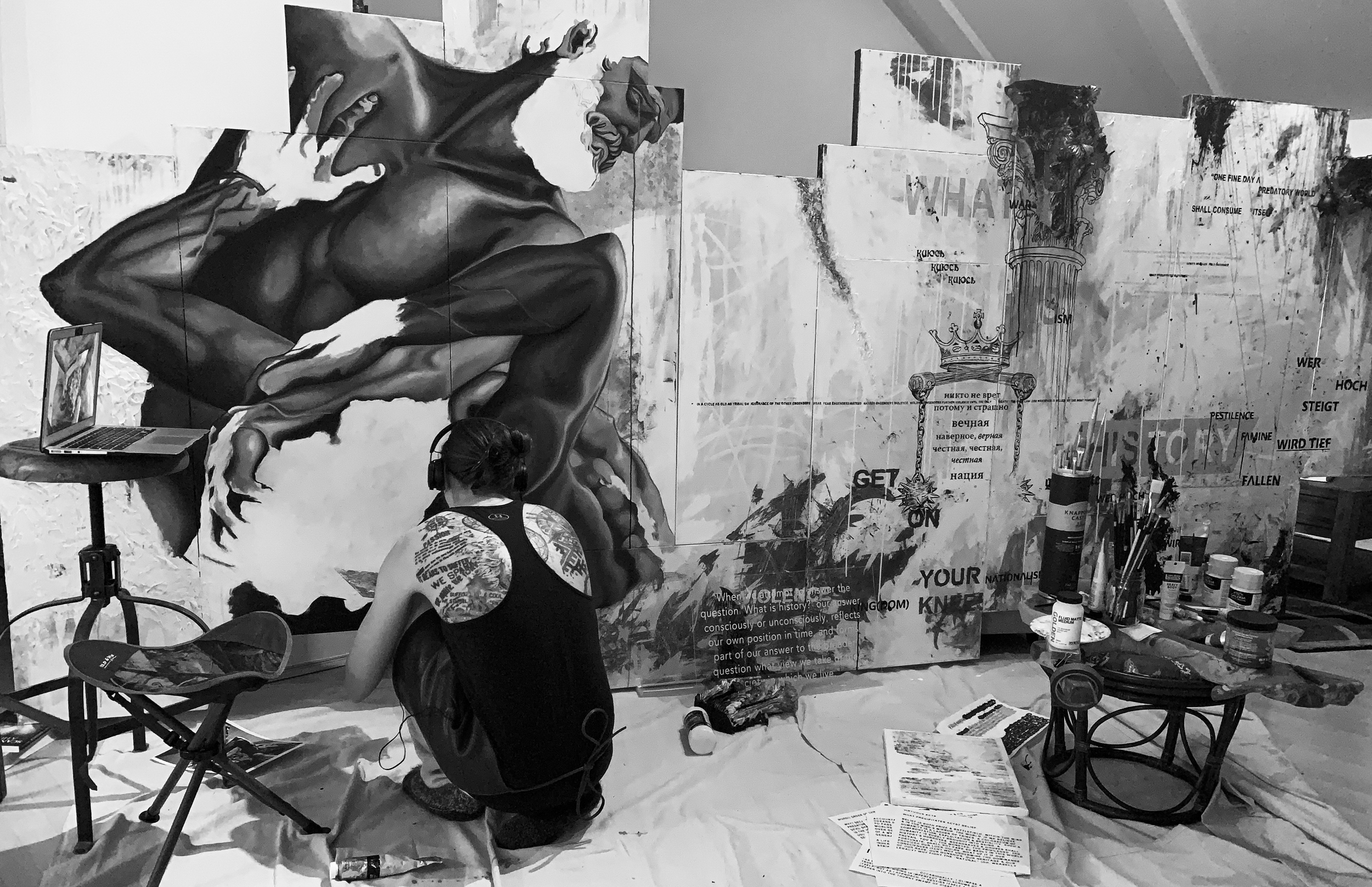 Black and white photo of a person with tattoos on their arm sitting on the floor, looking at a large artist's canvas, with art supplies and a laptop nearby, in an art studio or gallery setting.