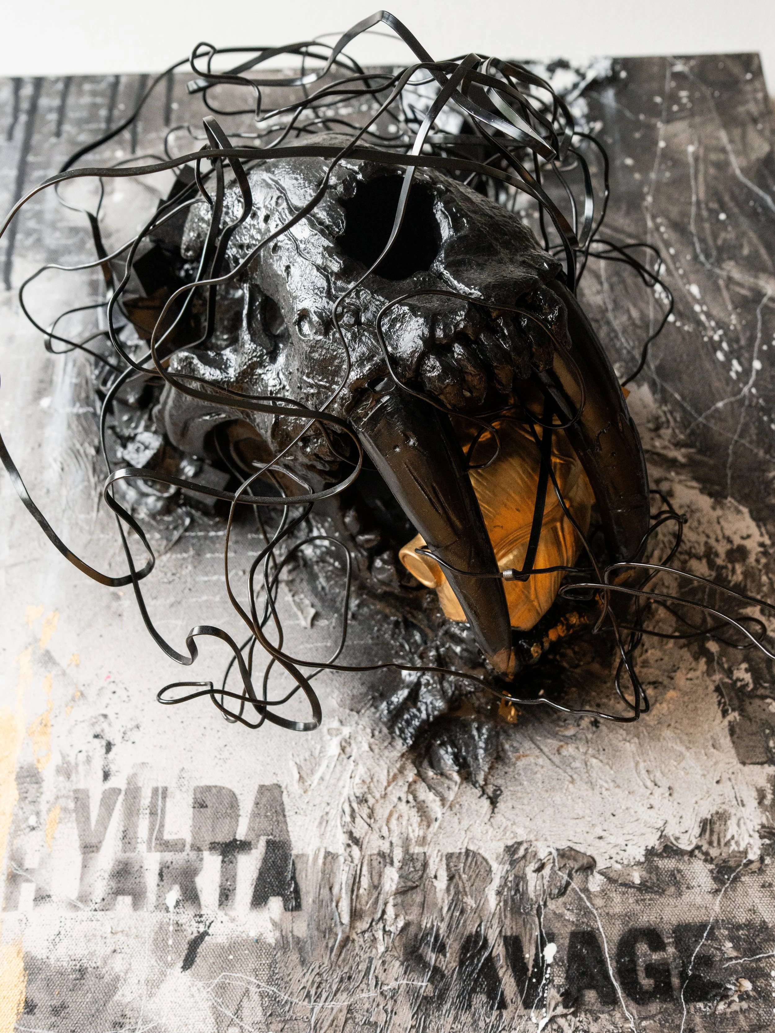 Close-up of a metallic skull sculpture with black wires tangled on top, placed on a printed surface with black and white text and graphics.