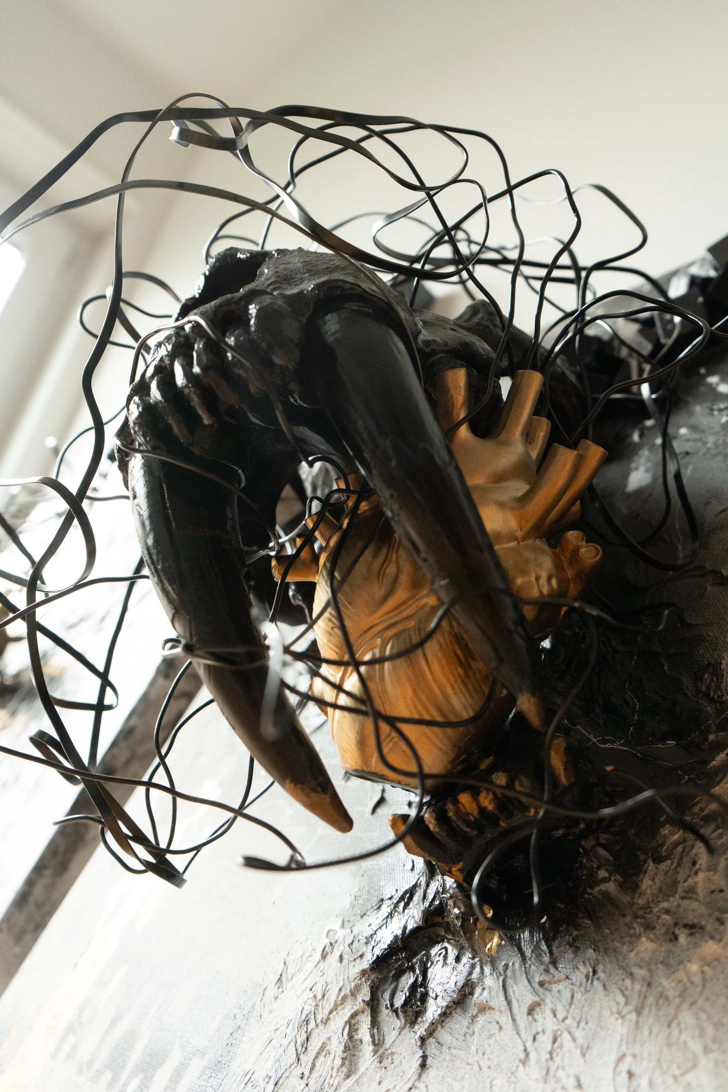 Close-up of a wooden sculpture with black wire accents and melted dark metal around it on a white surface.