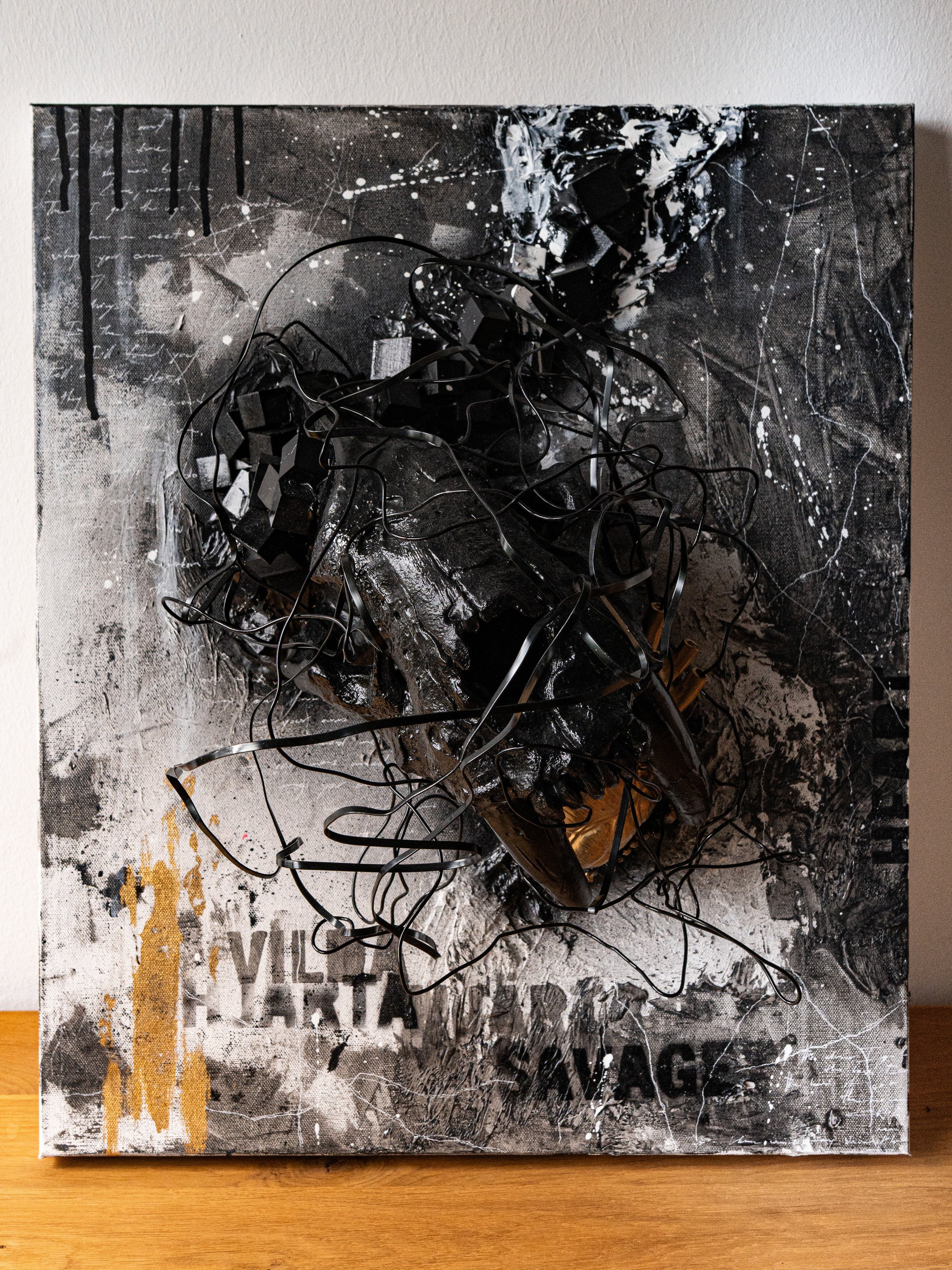 Mixed media artwork featuring black, white, and gold paint splatters, drips, scribbles, and tangled black wires or strips on a canvas with a collage background and partially visible text.