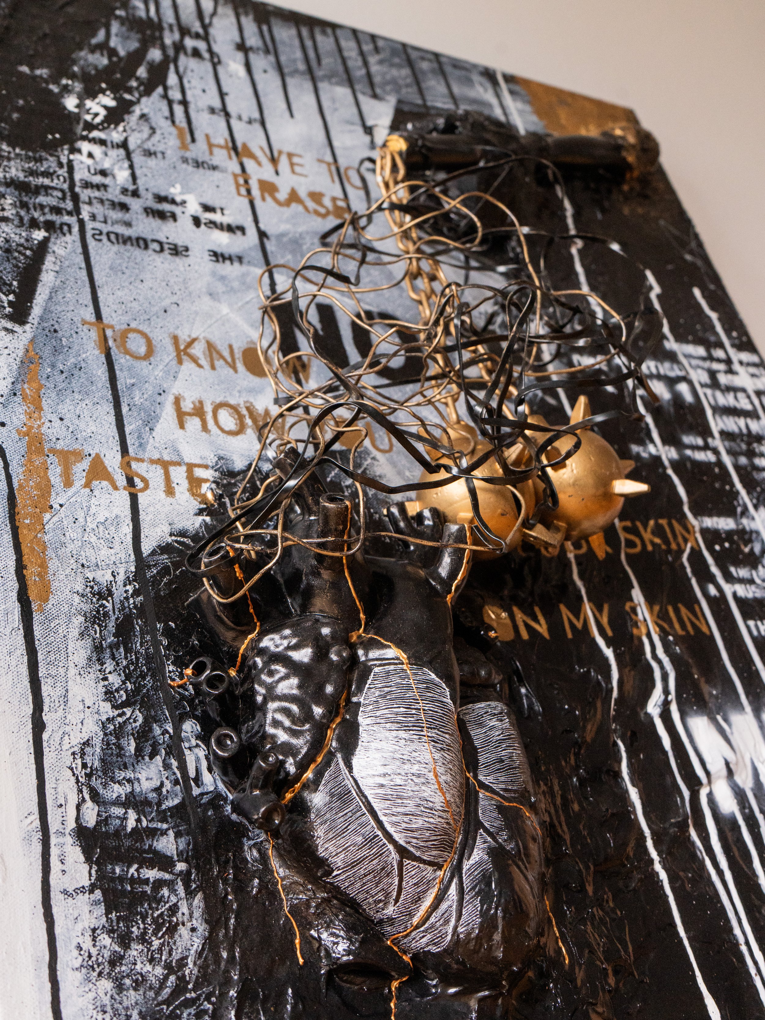 Mixed media art piece with metallic wires, gold-colored shapes, and abstract text and images, predominantly in black, white, and gold, with layered textures.