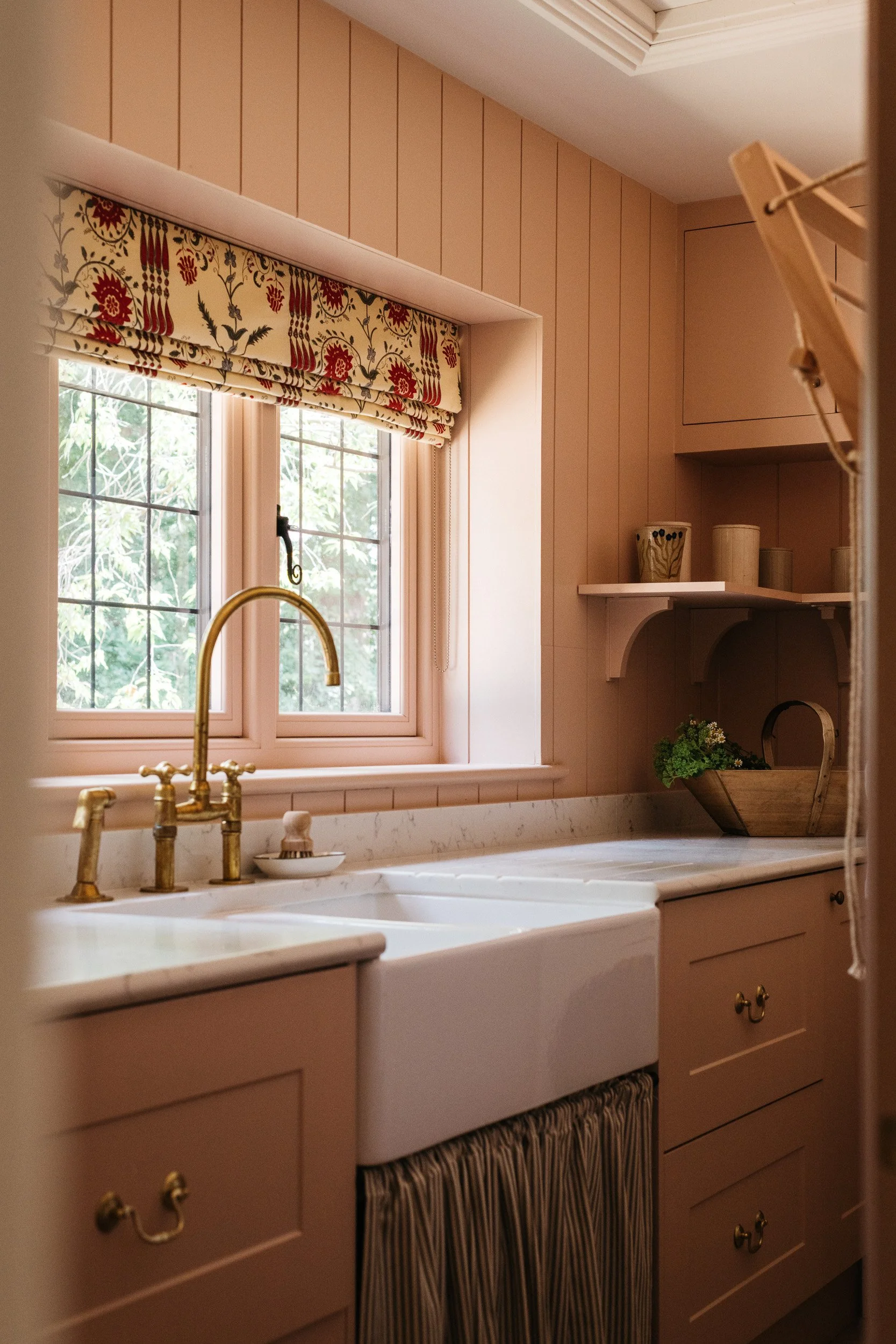 A kitchen window with a floral valance above it, a brass faucet over a white marble sink, and beige cabinetry with gold knobs, a shelf with decorative containers, and a basket with greenery.