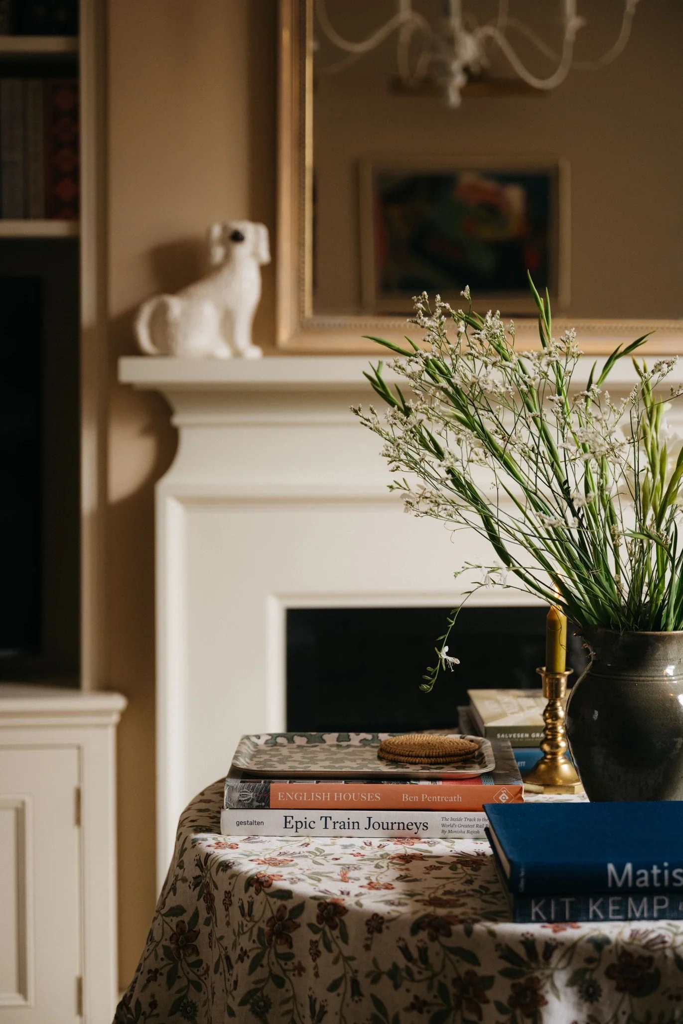 Living room with a floral tablecloth, books including 'English Houses' by Ben Pentreath and 'Epic Train Journeys', a black vase with greenery and white flowers, a gold candlestick, a large mirror, framed art on the wall, and a decorative dog figurine