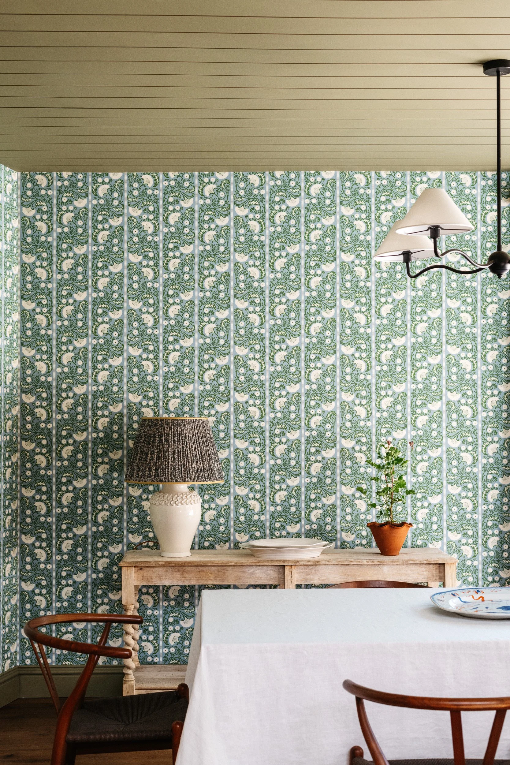 Interior of a dining room with a table covered with a white tablecloth, surrounded by wooden chairs, a wooden side table with a white ceramic lamp with a dark lampshade, some plates, a small green potted plant, and patterned wallpaper with green and 