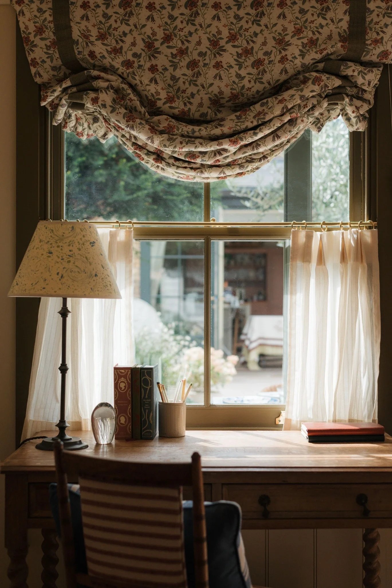 A cozy home office scene with a wooden desk, a lamp, books, and pens. There is a window with floral curtains letting in natural light, and a view of a garden outside.