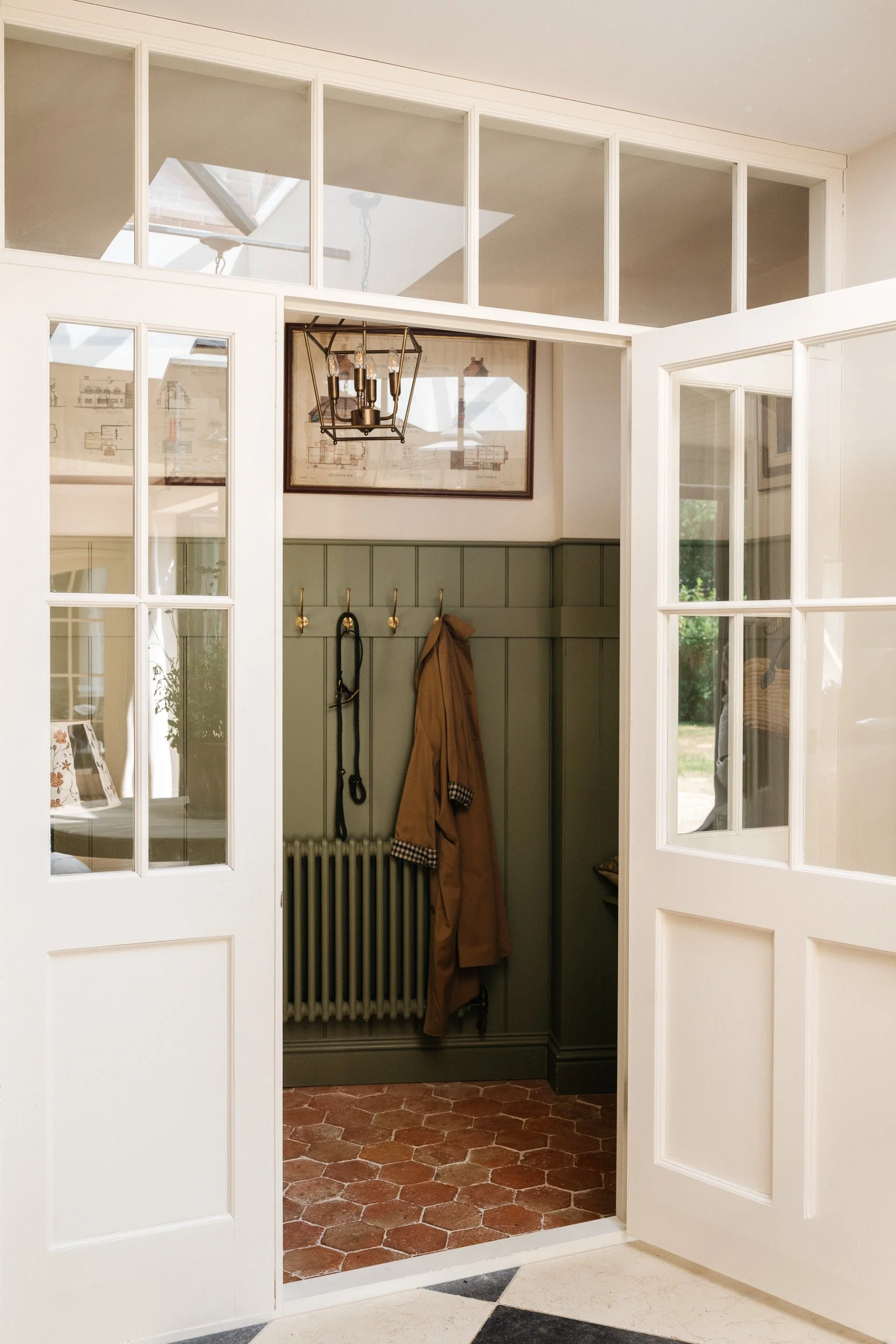 Interior view of a house entryway with white framed glass doors leading to a room with green wall paneling, brass hooks with coats, a framed blueprint on the wall, and a vintage radiator, with terracotta tile flooring.