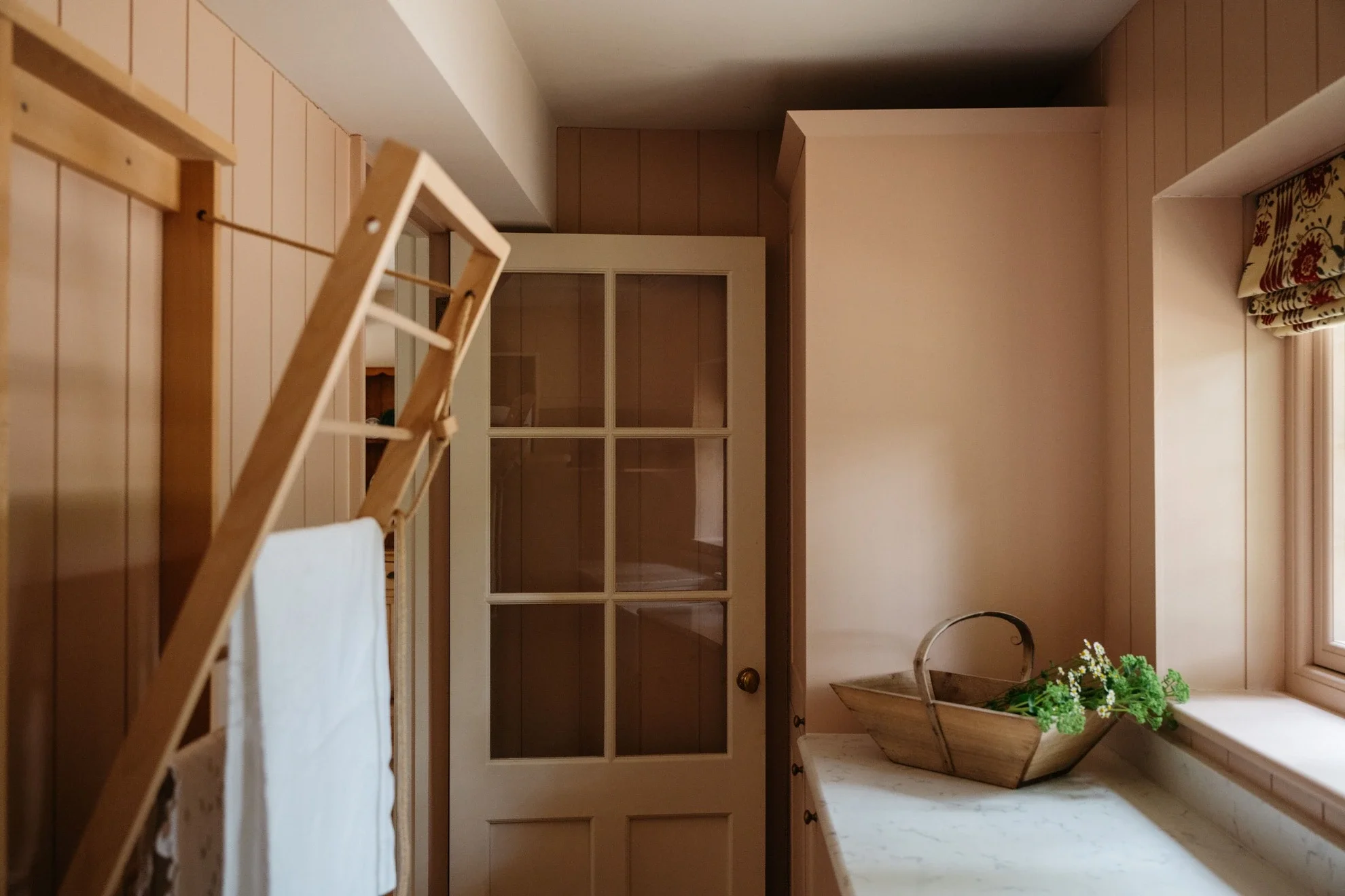 A cozy kitchen with wooden paneling, a window with floral curtains, and a countertop with a basket of green plants.