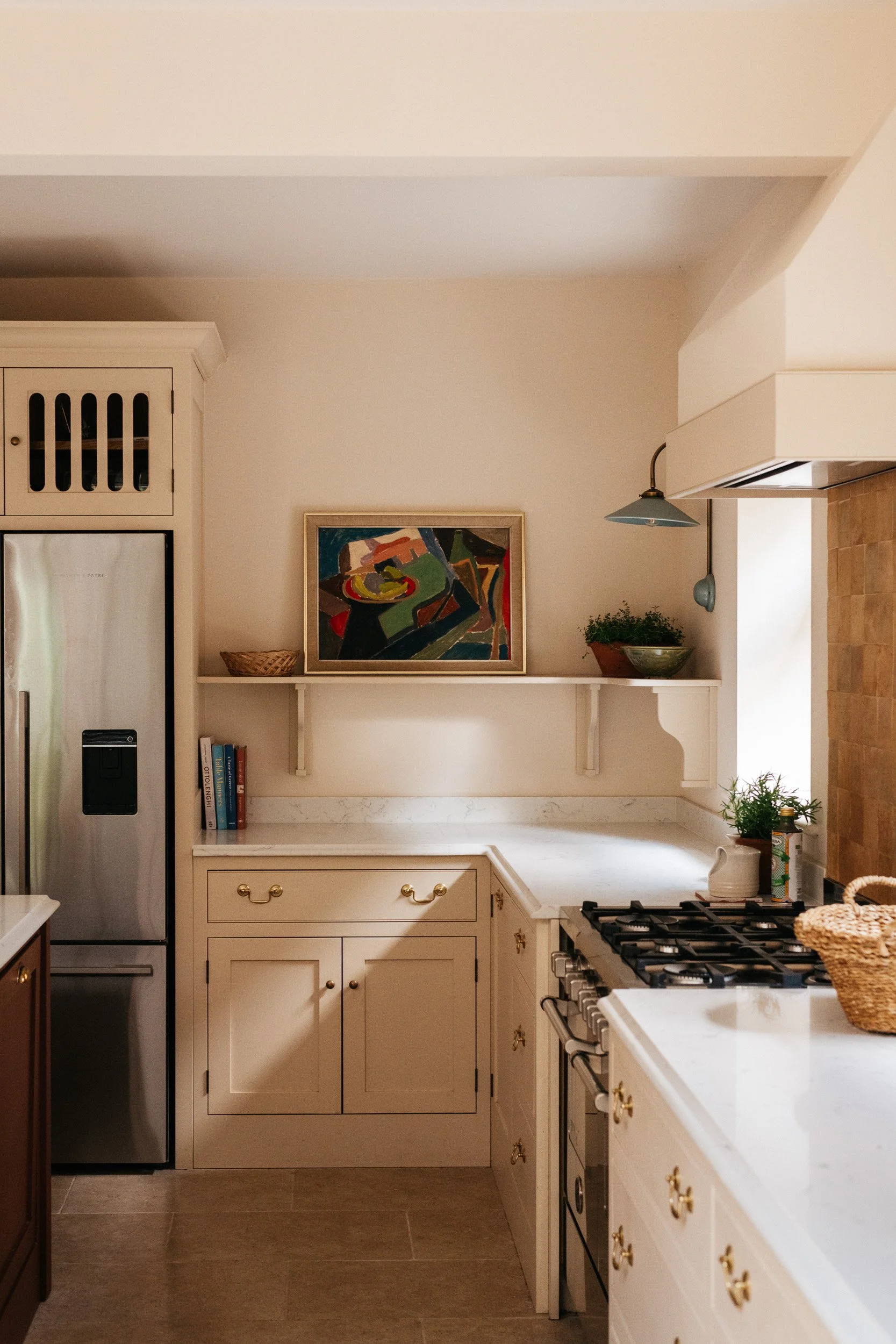 Bright kitchen with cream-colored cabinets, a stainless steel refrigerator, a colorful abstract painting on the wall, a white marble countertop, a gas stove, and decorative plants.