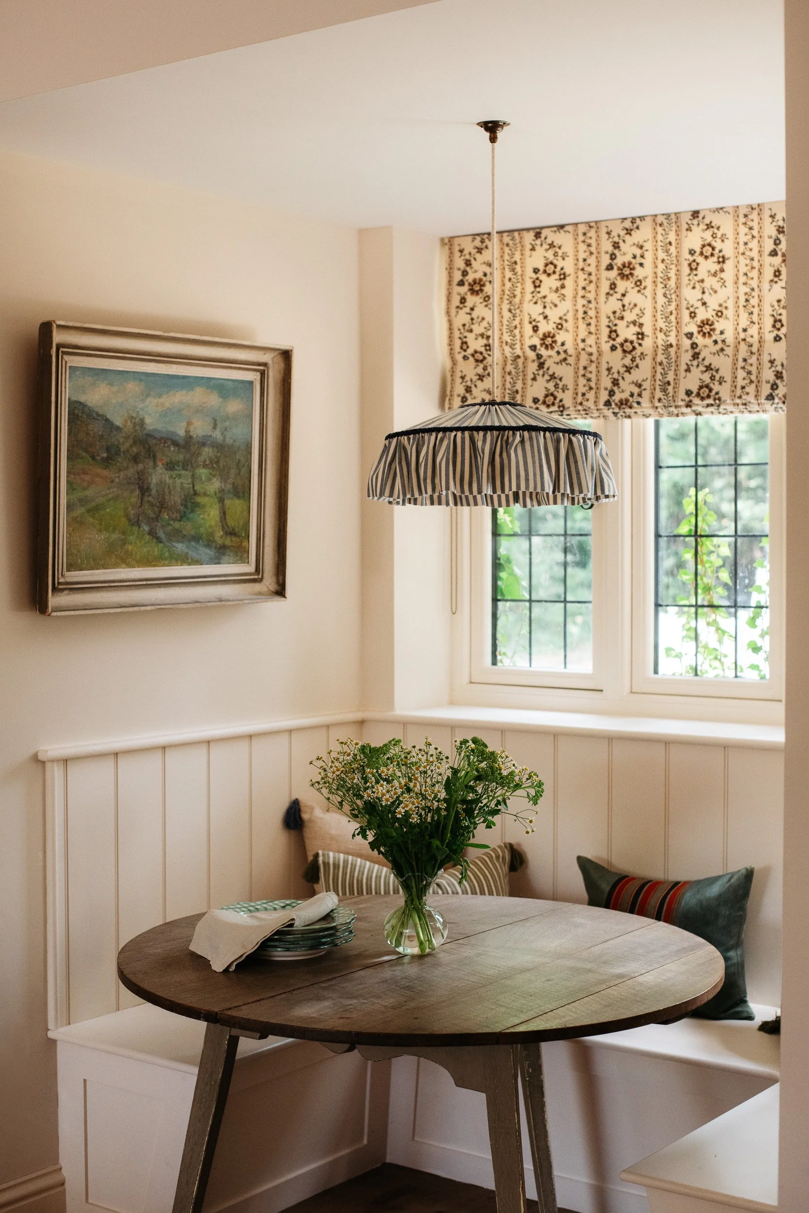 A cozy dining area with a round wooden table, a vase of wildflowers, and a window with floral Roman shades. A framed landscape painting hangs on the wall, and a striped fabric pendant light hangs from the ceiling.