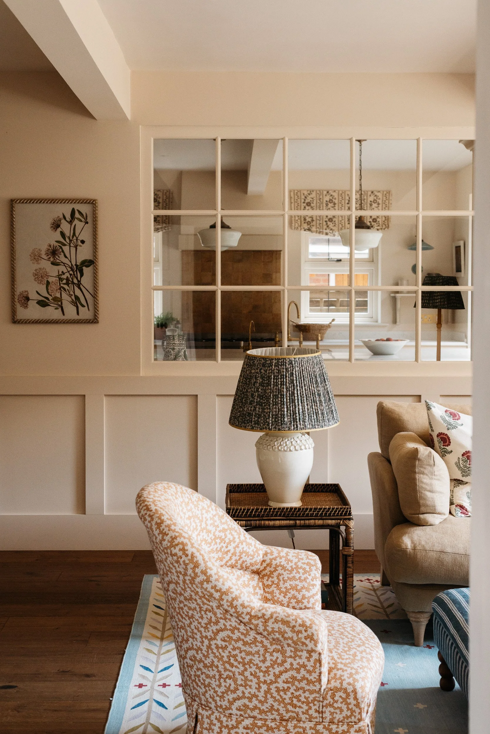A cozy living room with a patterned armchair in the foreground, a large beige sofa with floral pillows, and a table lamp on a wicker side table. Behind the sofa is a painted wall and a large mirror, reflecting a kitchen area with a window, fruit bowl