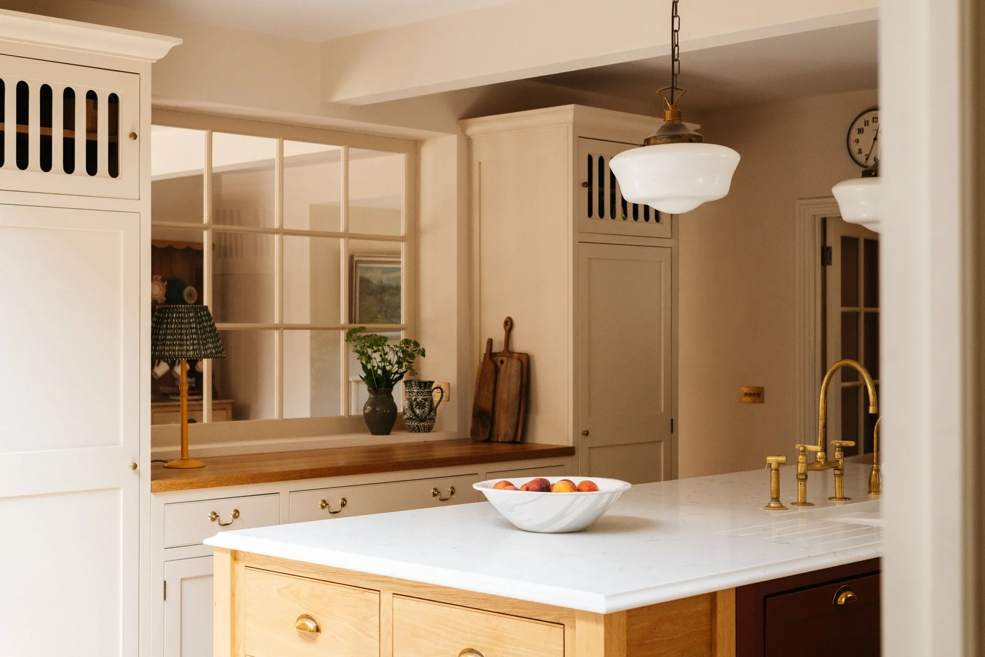 A kitchen with white cabinets, a white countertop, and a white island. There is a brass faucet and sink, a bowl of peaches on the island, and a window with a kitchen view. Two hanging light fixtures are visible, along with a clock and framed artwork 