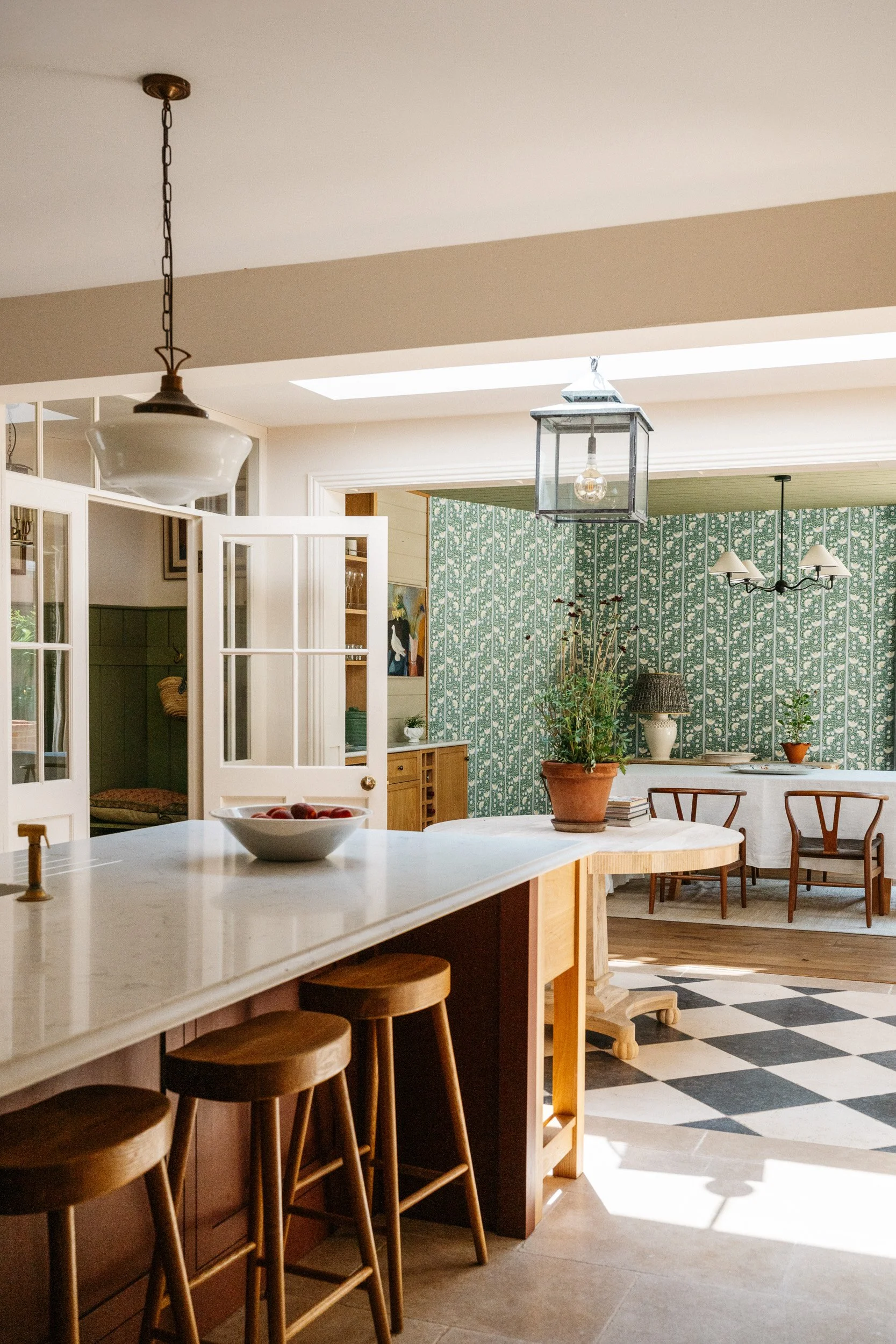 A bright, well-lit kitchen with white cabinetry, a marble island with wooden stools, and a patterned green wallpapered wall with decorative plants and artwork.